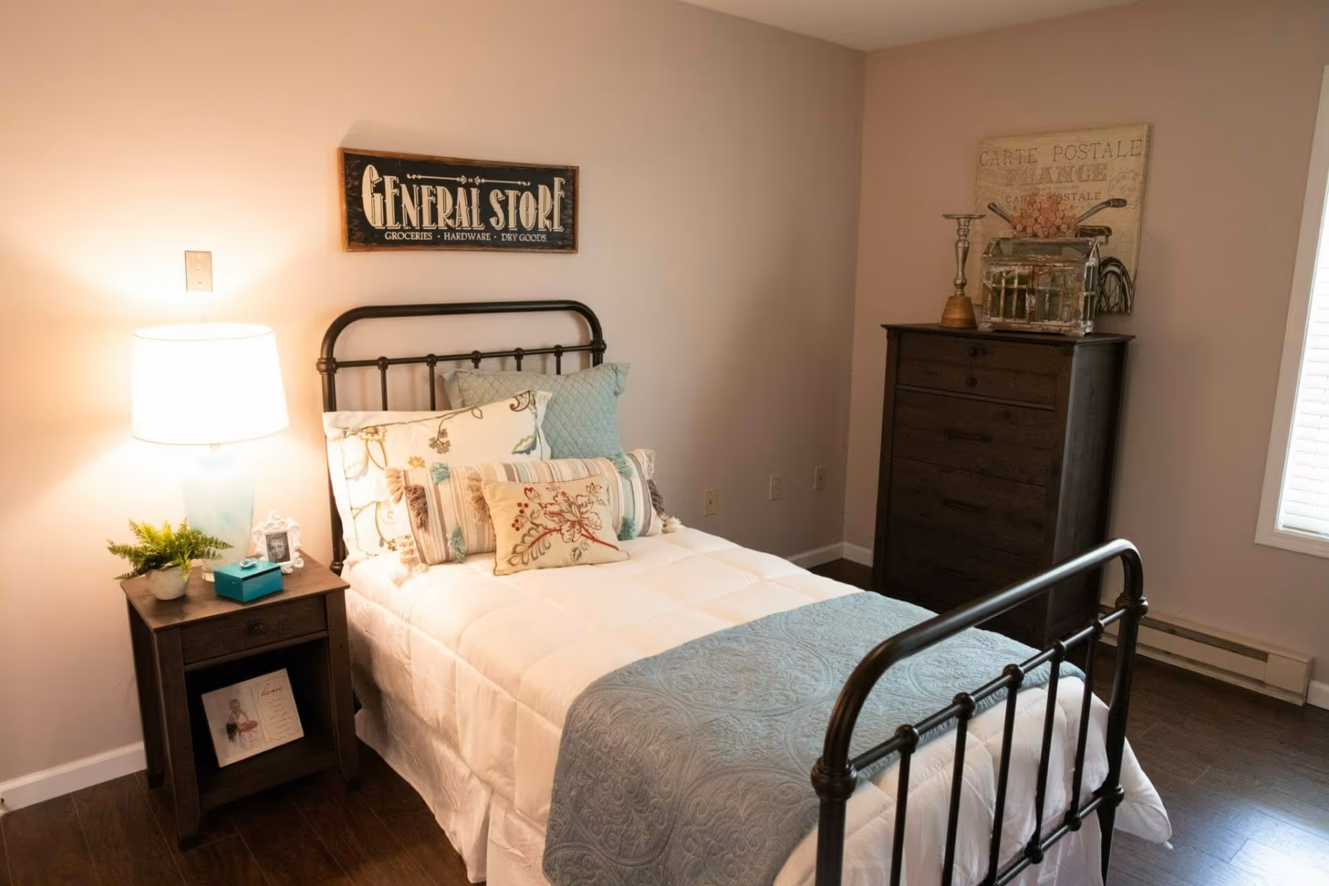A cozy bedroom with a single bed featuring a black metal frame, white bedding, and multiple decorative pillows. A wooden nightstand with a lamp, a small plant, a tissue box, and a framed photo sits next to the bed. On the wall above the bed is a sign that reads 'GENERAL STORE Groceries Hardware Dry Goods.' Across from the bed is a dark wooden dresser with decorative items on top and a framed artwork on the wall behind it. The room has wooden flooring and a window with blinds.