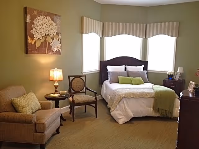 A cozy bedroom with a bed featuring white and green bedding, two pillows, and a green throw blanket. There is a wooden chair and a cushioned armchair with a green pillow next to a small round table with a lamp and a cup. The room has three windows with striped valances and a floral painting on the wall.