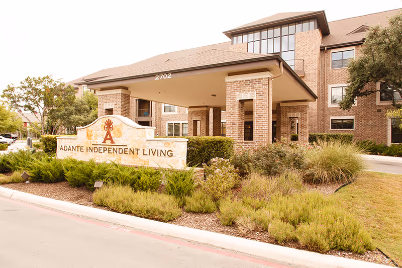 Exterior view of Adante Independent Living facility showing a brick building with multiple windows, a covered entrance supported by brick columns, and a stone sign with the facility name surrounded by landscaped greenery.