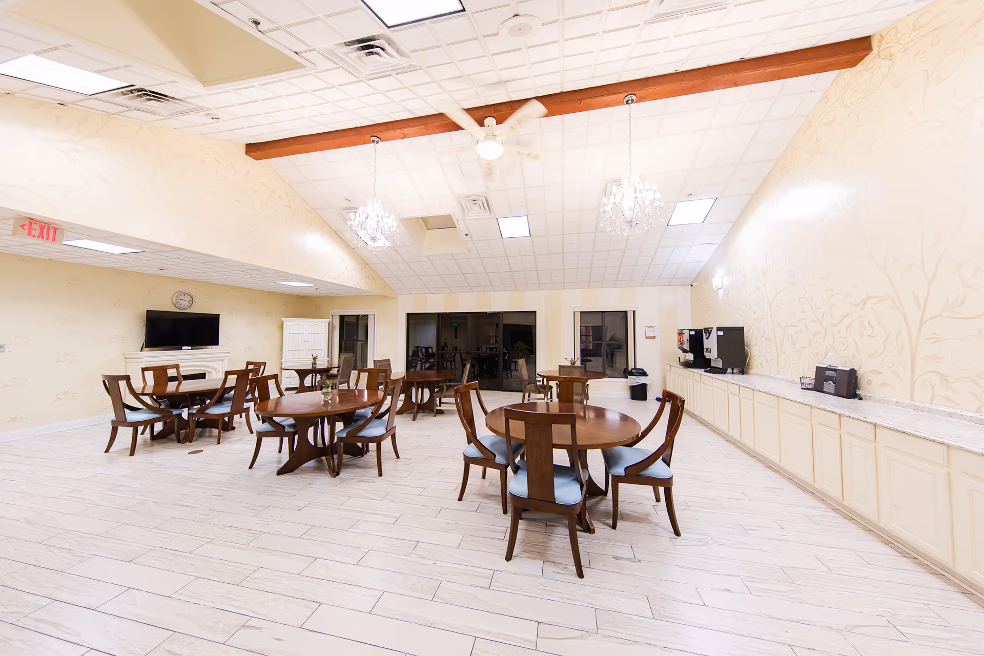 A spacious dining room with several round wooden tables and chairs arranged neatly. The room has light-colored walls with subtle floral patterns, a tiled floor, and a high ceiling with wooden beams and chandeliers. There is a TV mounted on the wall above a white fireplace, and a long counter with coffee machines and other appliances on the right side. Large windows and glass doors are visible at the far end of the room.