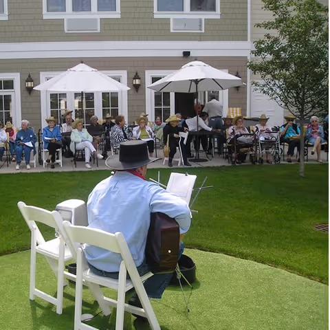 A man wearing a black hat and light blue shirt is playing guitar and singing outdoors on a grassy area in front of a seated audience of elderly people. The audience is sitting on chairs under white umbrellas outside a building with beige siding and multiple windows. Some audience members are using walkers or wheelchairs.