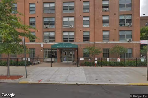 Brick senior living building with green awning entrance, trees, benches, and fenced walkway.