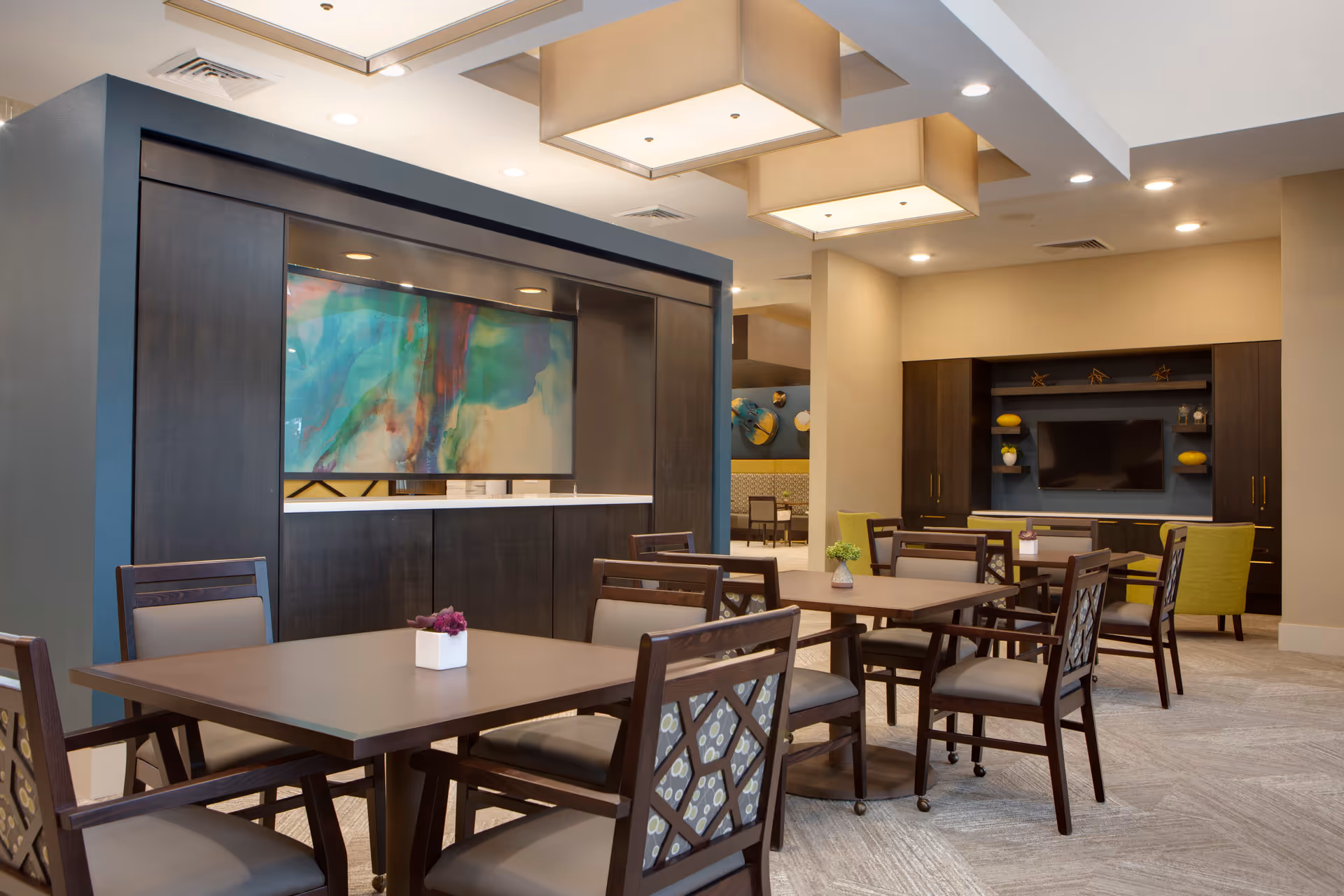 A modern senior living common area with multiple wooden tables and chairs arranged for seating. The room features a large abstract painting on a dark wood partition, recessed ceiling lights, and a built-in entertainment center with a flat-screen TV and decorative items. The space is well-lit and decorated with neutral tones and pops of color from cushions and decor.