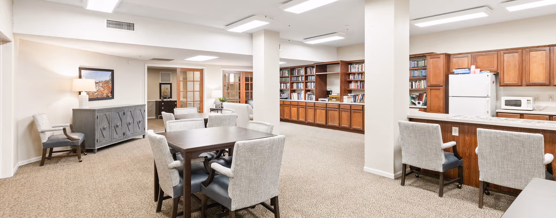 A bright and spacious common area in a senior living facility featuring a dining table with six upholstered chairs, a kitchen area with wooden cabinets, a white refrigerator, and a microwave. There is a large built-in wooden bookshelf filled with books along the wall. The room has beige carpet flooring, white walls, and ceiling lights. A sideboard with a lamp and a framed picture is visible on the left side.
