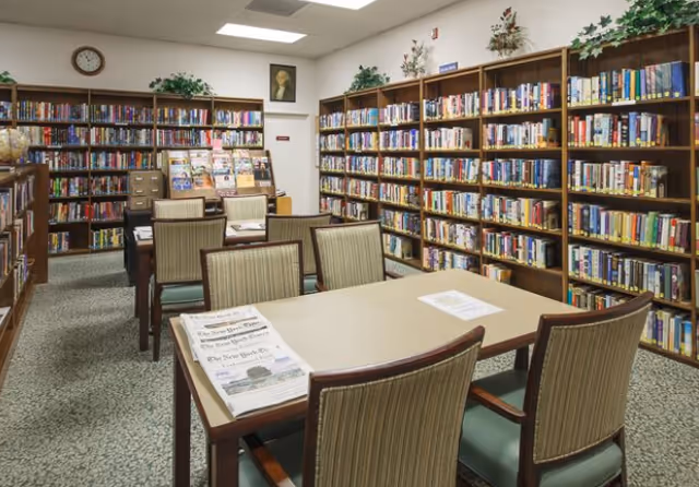 Interior view of a library room with multiple wooden bookshelves filled with books along the walls. Several tables with chairs are arranged in the center of the room, some with newspapers and papers on them. The room has a carpeted floor, a clock on the wall, and decorative plants on top of the bookshelves.