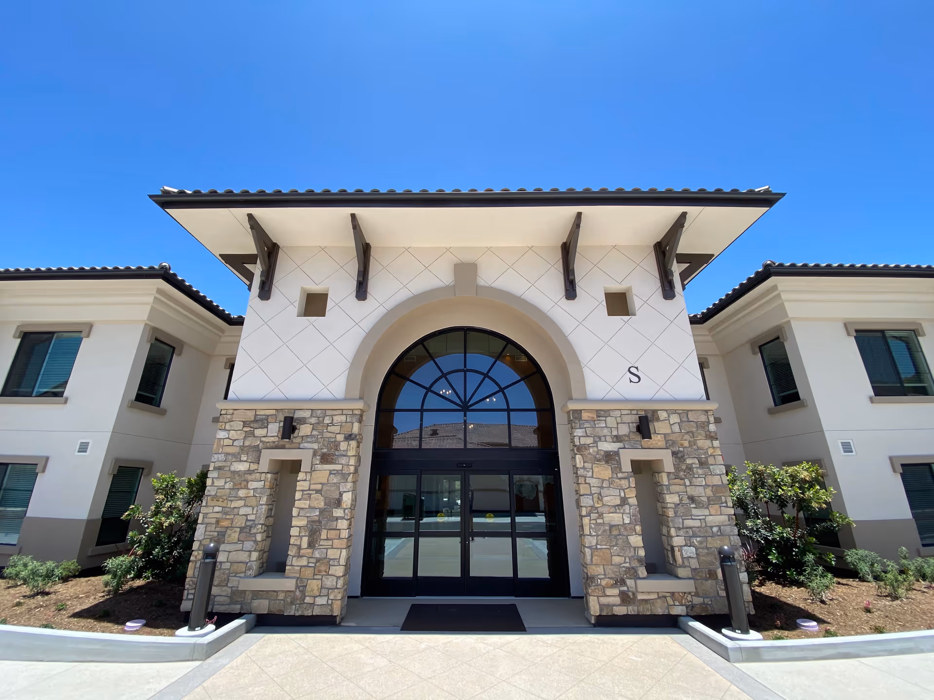 Front exterior view of a building with a large arched glass entrance door, stone and stucco facade, and a clear blue sky above.