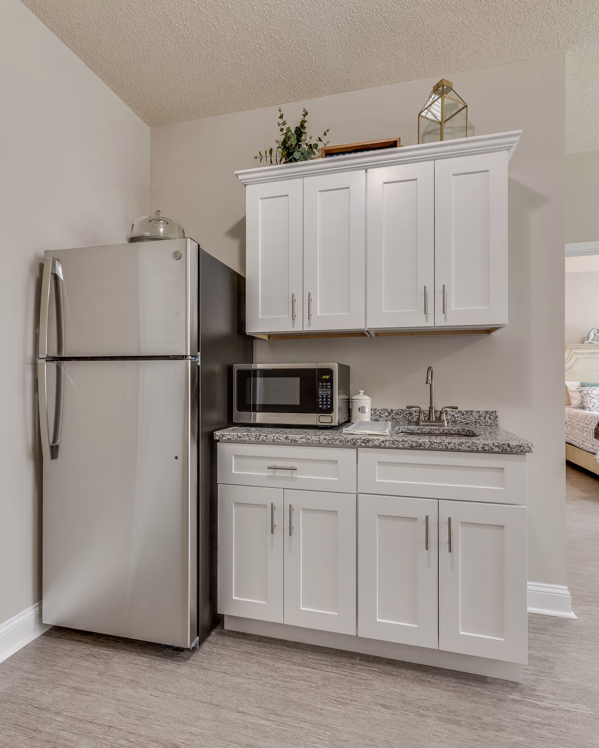 A small kitchen area with white cabinets, a granite countertop, a stainless steel refrigerator, a microwave, and a sink. There are decorative items on top of the upper cabinets, and a glimpse of a bedroom is visible through an open doorway.
