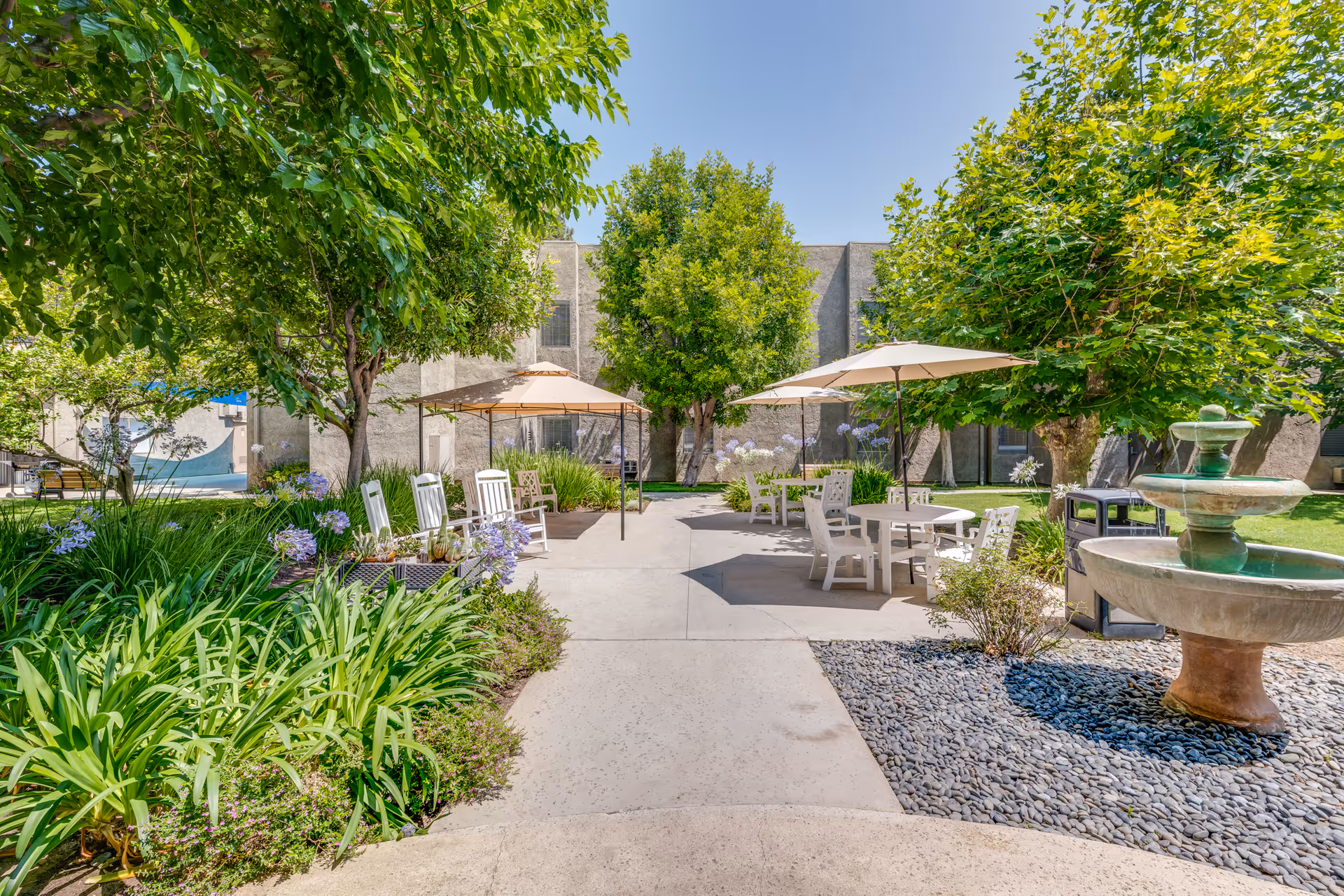 Outdoor courtyard area with concrete pathways, green trees, flowering plants, white patio tables and chairs with umbrellas, and a stone water fountain on the right side under a clear blue sky.