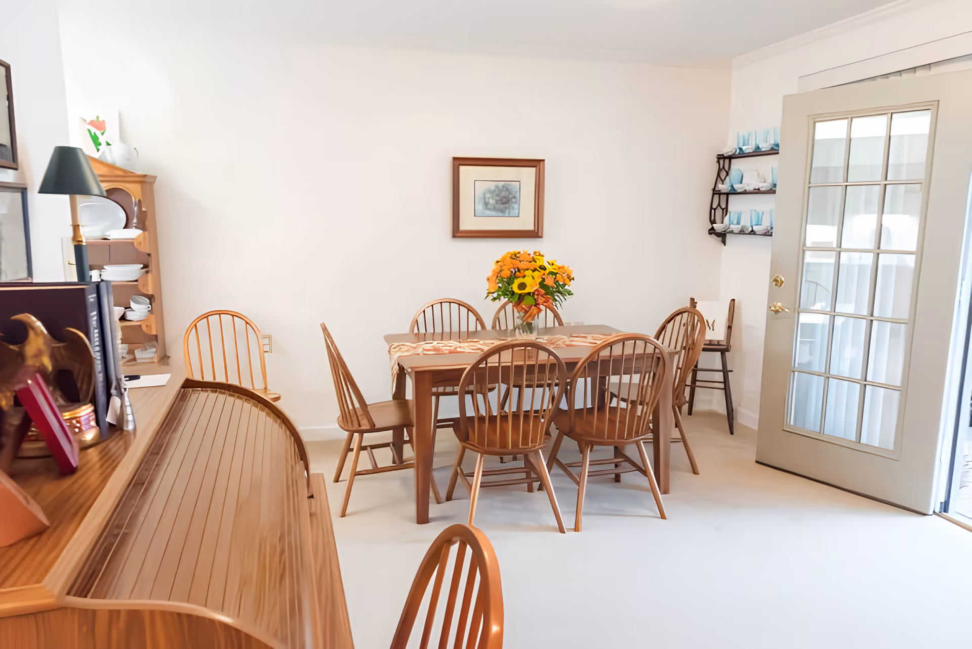 Bright dining room with a wooden table surrounded by chairs, a vase of flowers on the table, shelving and a glass-paneled door.