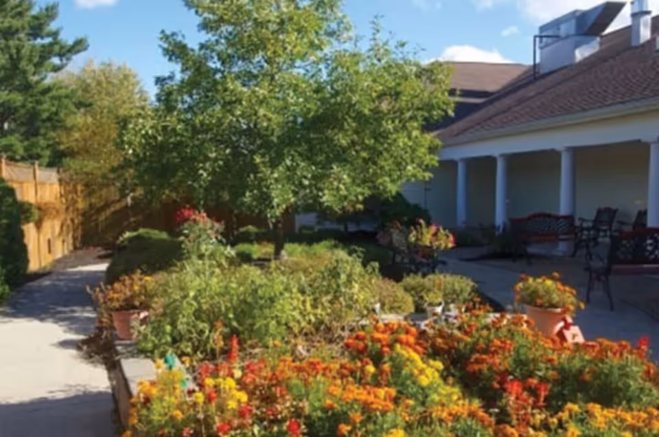 A vibrant garden area with colorful flowers and green shrubs in front of a building with a covered porch. The porch has several benches and chairs, and there is a paved walkway leading through the garden. Trees and a wooden fence are visible in the background under a partly cloudy sky.