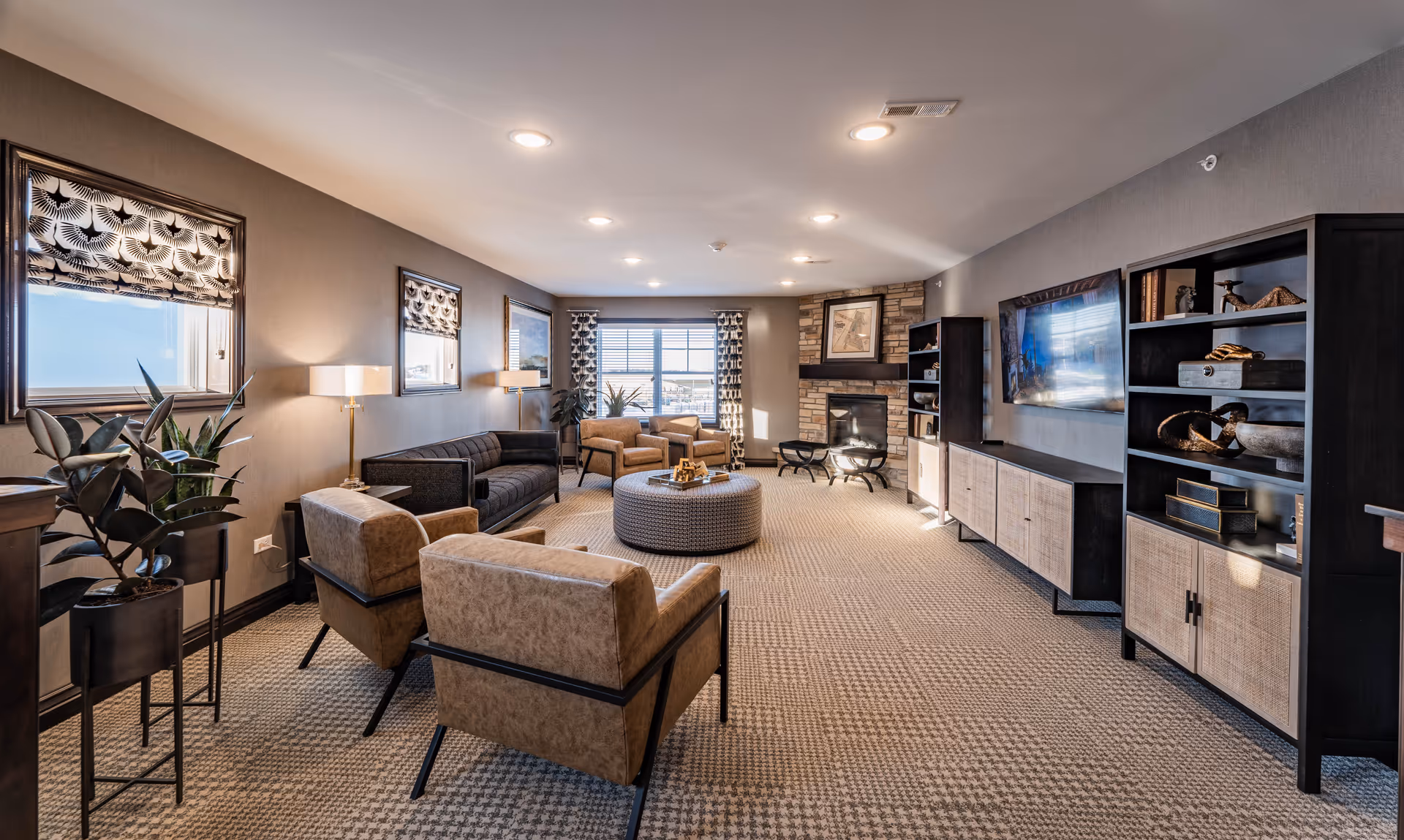 A spacious and well-lit senior living common area featuring a patterned carpet, two tan armchairs, a dark sofa, a round woven ottoman coffee table, two additional armchairs near a large window with patterned curtains, a stone fireplace with framed artwork above it, and dark wood shelving units with decorative items and a mounted flat-screen TV.