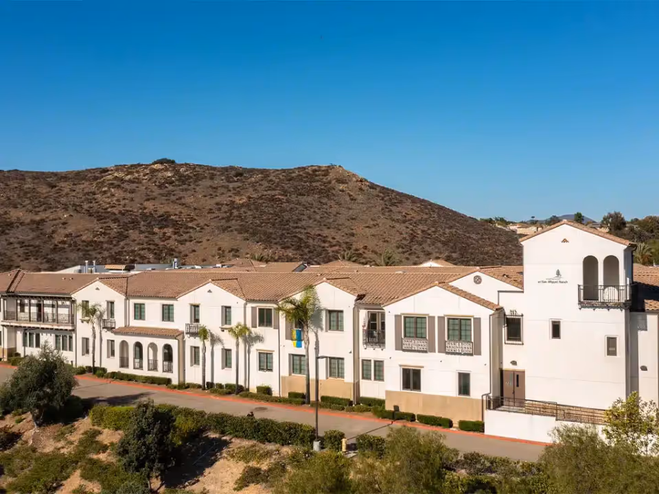 Exterior view of Westmont at San Miguel Ranch, a large white building with multiple windows and balconies, set against a backdrop of a dry, rocky hill under a clear blue sky.