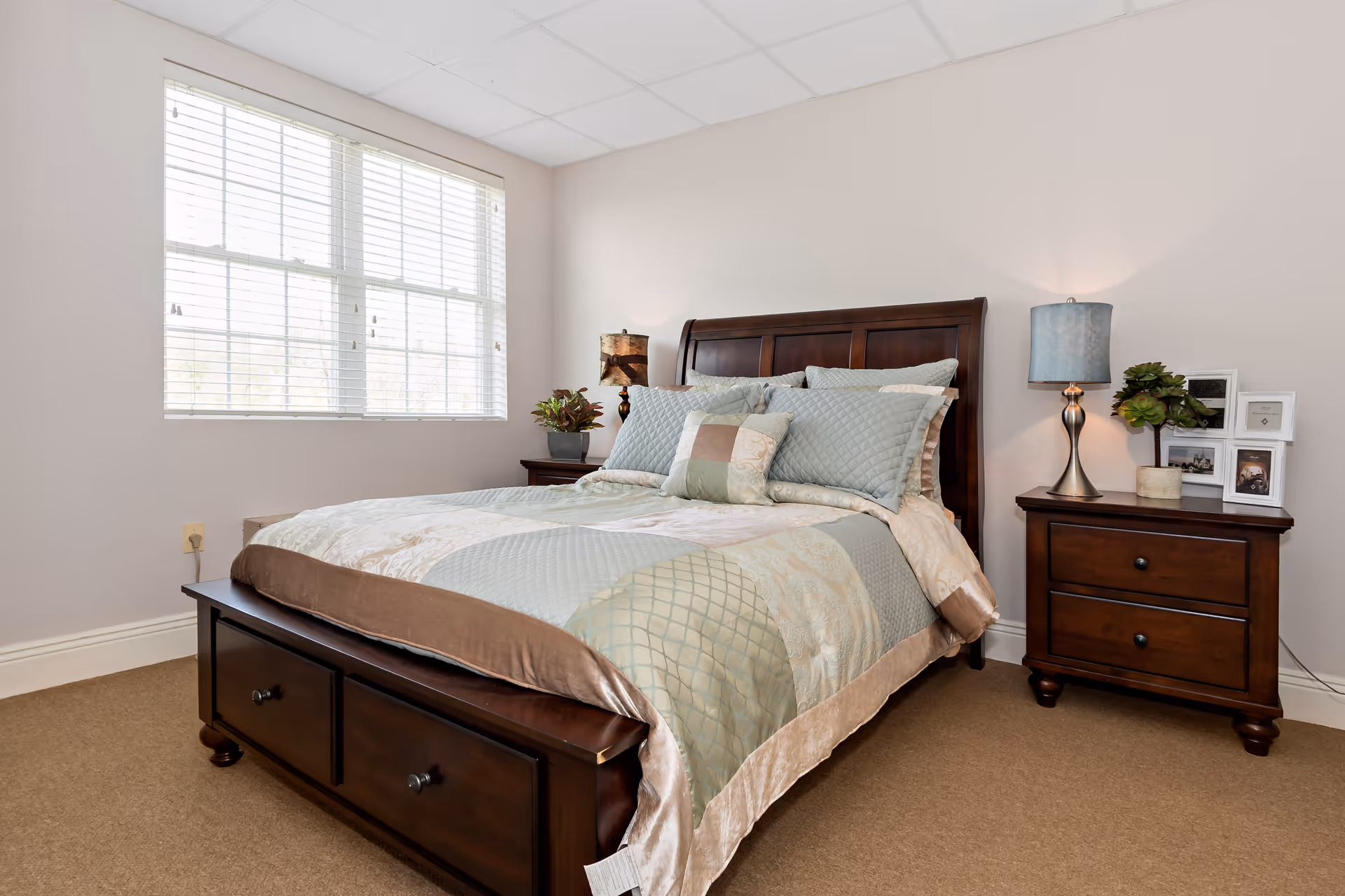 A neatly made bed with a wooden headboard and footboard in a bedroom. The bed has a light blue and beige quilt with multiple pillows. There are two wooden nightstands on either side of the bed, each with a lamp and decorative items including framed photos and a small plant. A large window with white blinds allows natural light into the room.