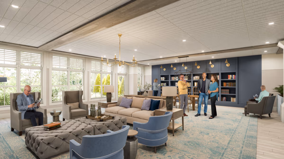 Bright, spacious communal living room with sofas, armchairs, a large ottoman, bookshelf wall, and several people socializing and reading.