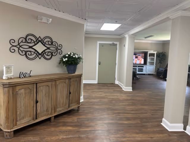 Interior view of a senior living facility hallway with wooden flooring and beige walls. On the left side, there is a wooden cabinet with decorative items including a potted plant, a small framed sign, and a 'blessed' sign. A decorative black metal wall art piece hangs above the cabinet. The hallway leads to a room with a television and seating area visible in the background.