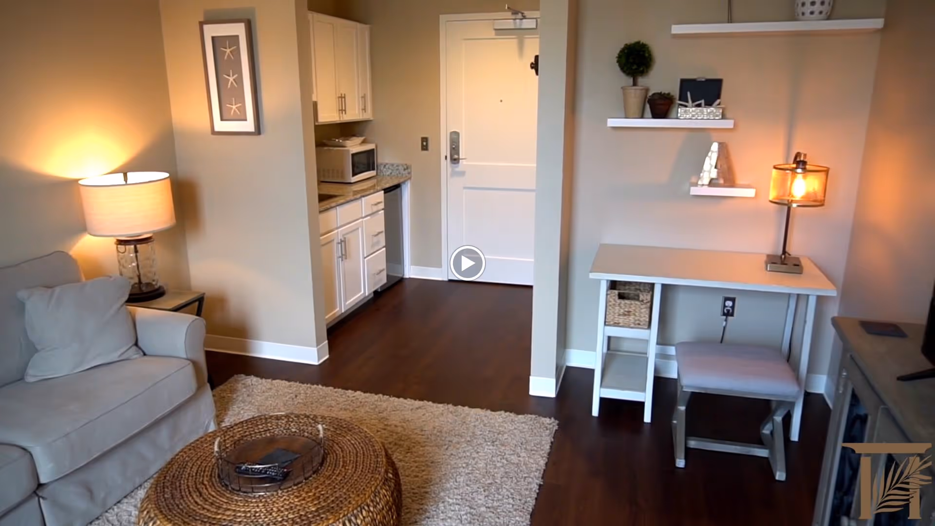 A cozy living area with a beige armchair, a round wicker coffee table on a light rug, a small kitchenette with white cabinets and a microwave, and a white desk with a lamp and decorative shelves on the wall. The floor is dark wood, and the walls are painted beige.