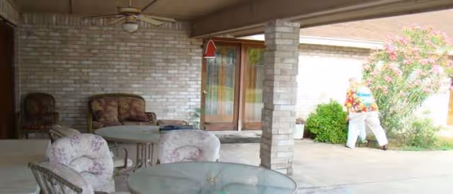 Outdoor patio with glass table and cushioned chairs, brick columns, sliding door, and a person walking.