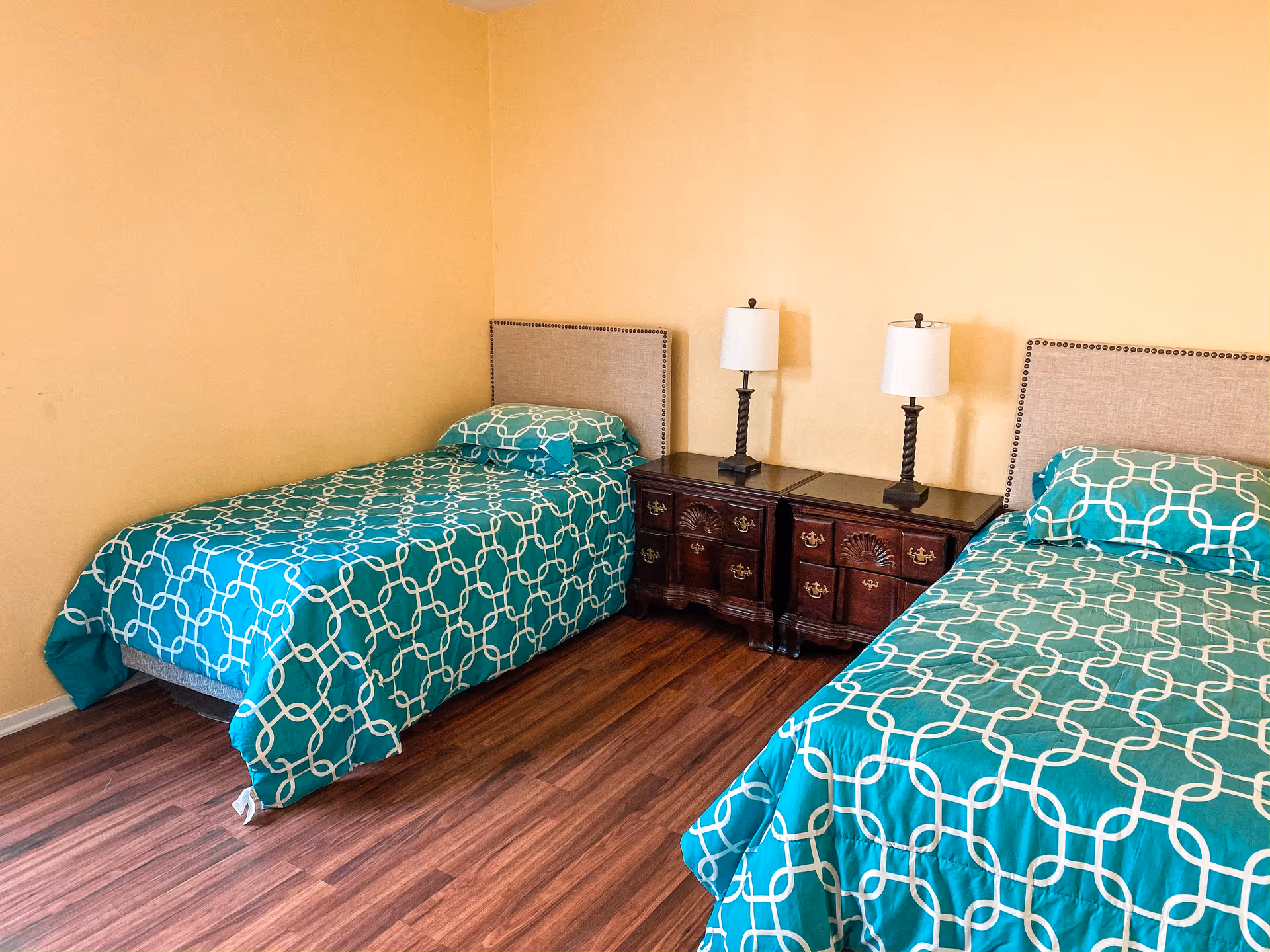 A bedroom with two twin beds featuring teal bedspreads with white geometric patterns. Each bed has a matching pillow and a beige upholstered headboard. Between the beds are two dark wooden nightstands with decorative carvings, each topped with a lamp with a white shade. The room has wooden flooring and light yellow walls.