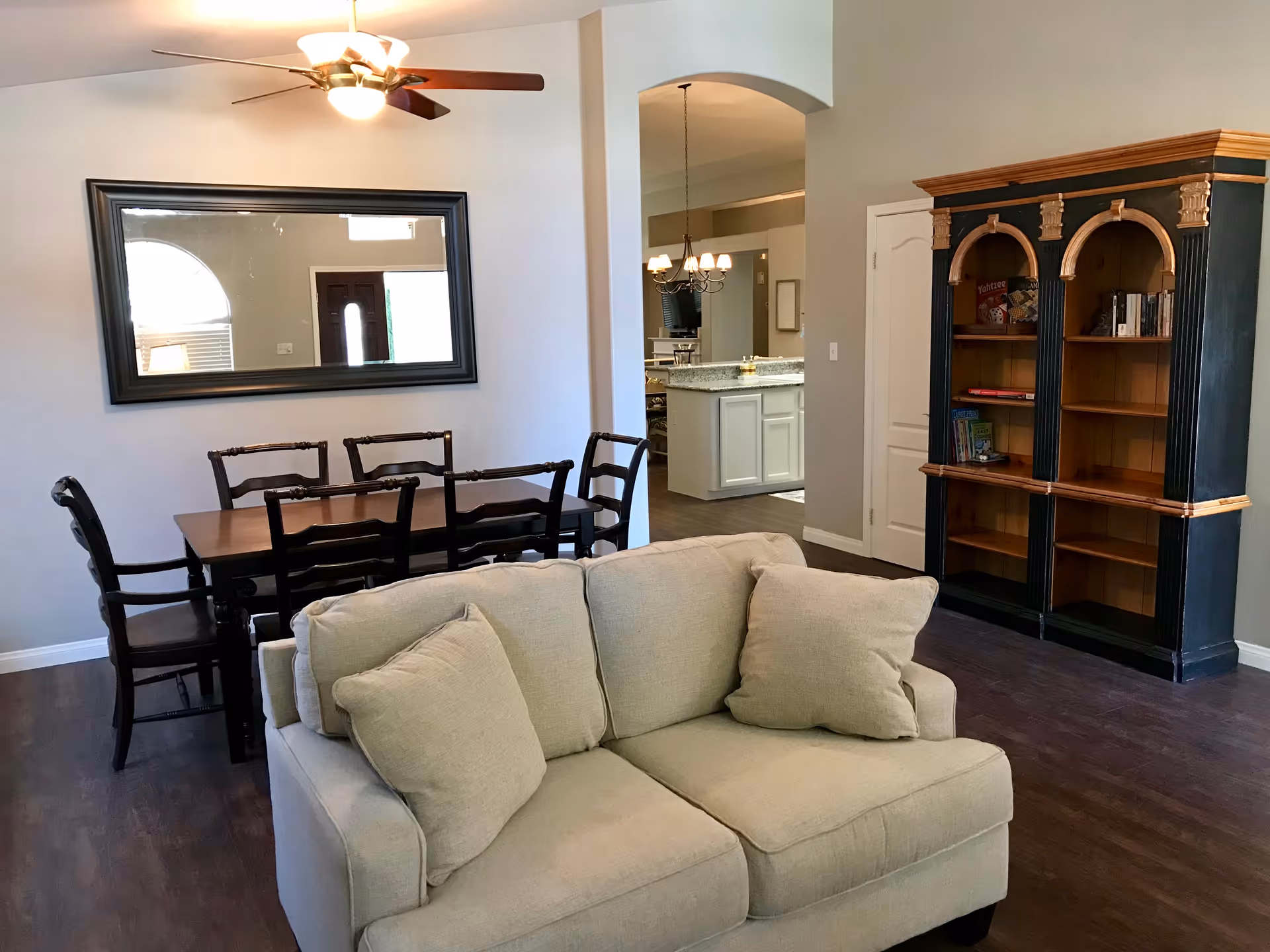 Interior view of a senior living facility showing a beige couch with two cushions in the foreground, a wooden dining table with six chairs against the wall, a large rectangular mirror above the dining table, a ceiling fan with lights, and a black and wood-toned bookshelf with books and games. In the background, a kitchen area with white cabinets and granite countertops is visible through an arched doorway.