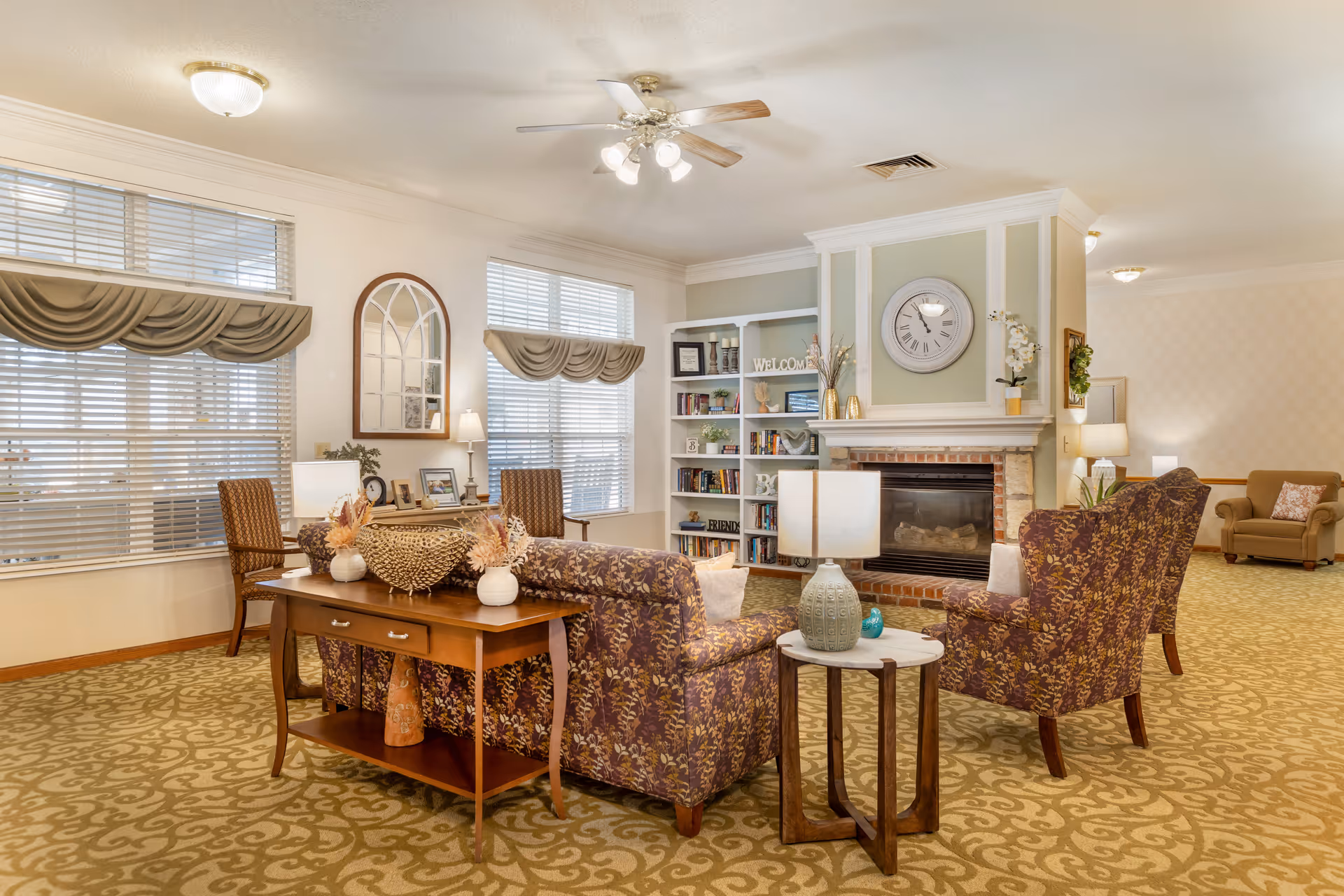 A cozy living room area in a senior living facility featuring patterned armchairs and a sofa arranged around a small round table with a lamp. Behind the sofa is a wooden console table with decorative vases. The room has large windows with blinds and valances, a ceiling fan with lights, a brick fireplace with a large clock above it, and built-in shelves filled with books and decorative items. The carpet has a floral pattern and the walls are painted in soft neutral tones.