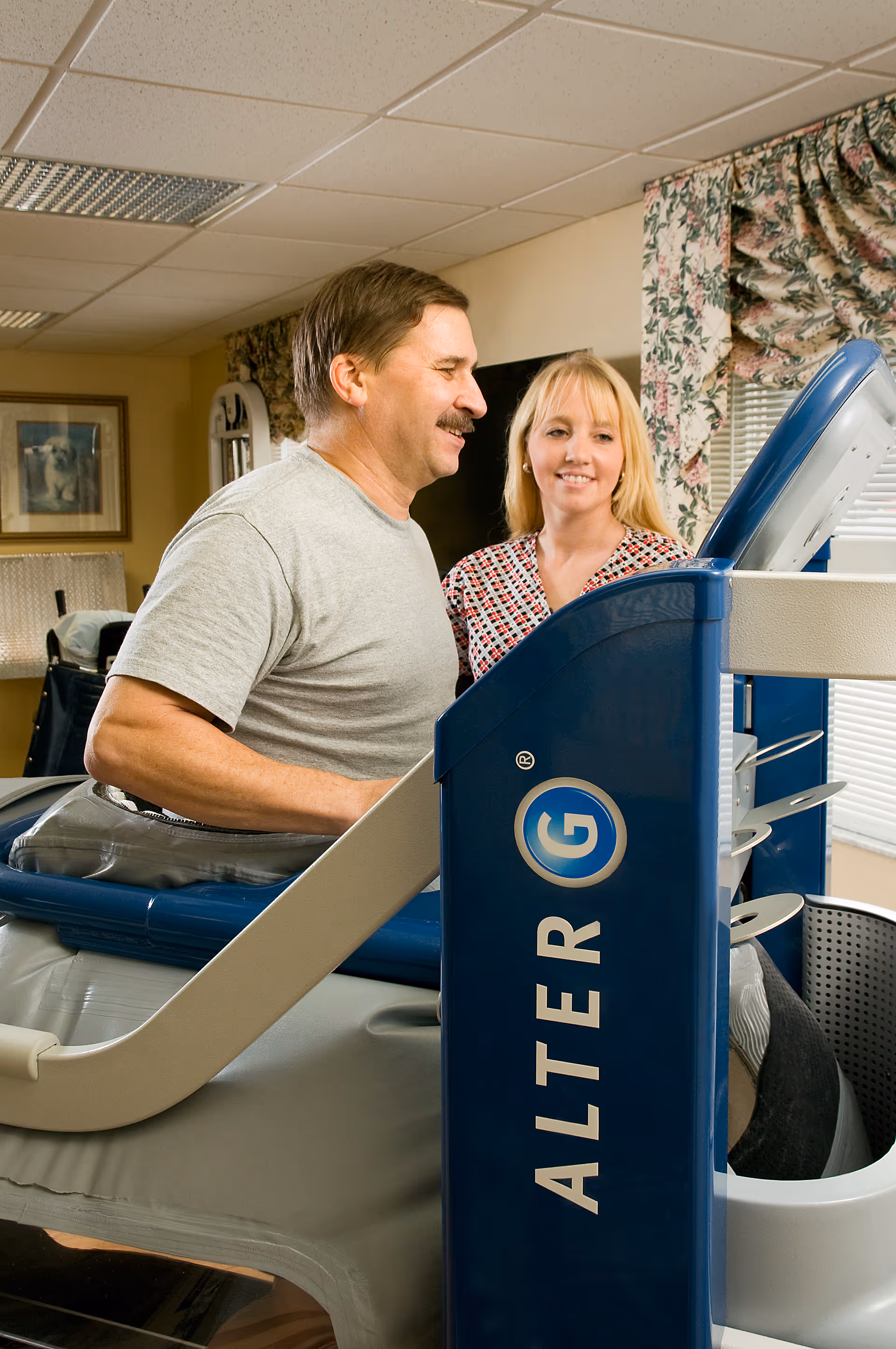 A man using an AlterG anti-gravity treadmill with the assistance of a woman in a room with floral curtains and framed artwork on the wall.