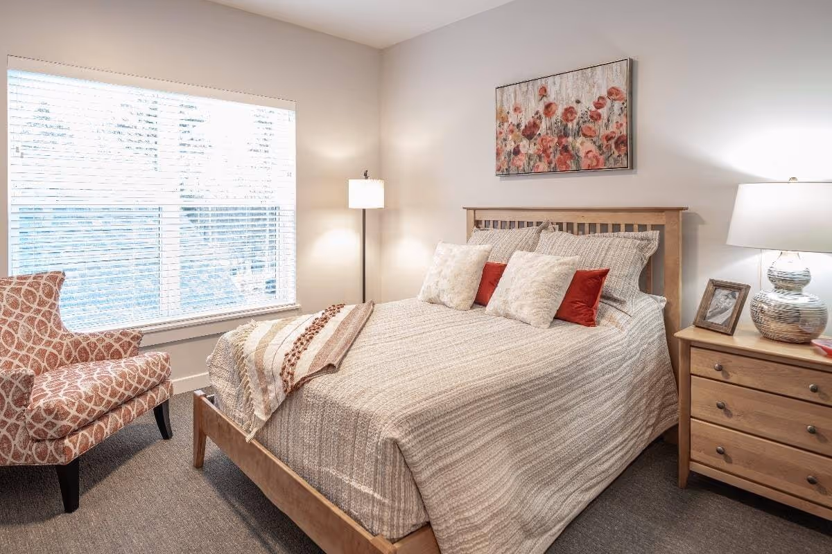 A cozy bedroom with a wooden bed frame, neatly made bed with beige and red pillows, a beige striped blanket, a wooden nightstand with a lamp and a framed photo, a patterned armchair near a large window with white blinds, and a floral painting on the wall.