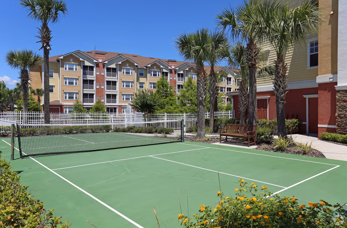 Outdoor tennis court with a net in the foreground, surrounded by palm trees and landscaping. In the background, there is a multi-story residential building with balconies under a clear blue sky.