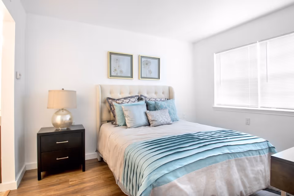 A bright bedroom with a neatly made bed featuring a tufted beige headboard, multiple decorative pillows in shades of blue and gray, and a blue-striped bedspread. There is a black nightstand with two drawers and a silver lamp with a beige lampshade on the left side of the bed. Two framed floral artworks hang on the white wall above the bed. A window with closed blinds is on the right side, allowing natural light to fill the room. The floor is wooden.