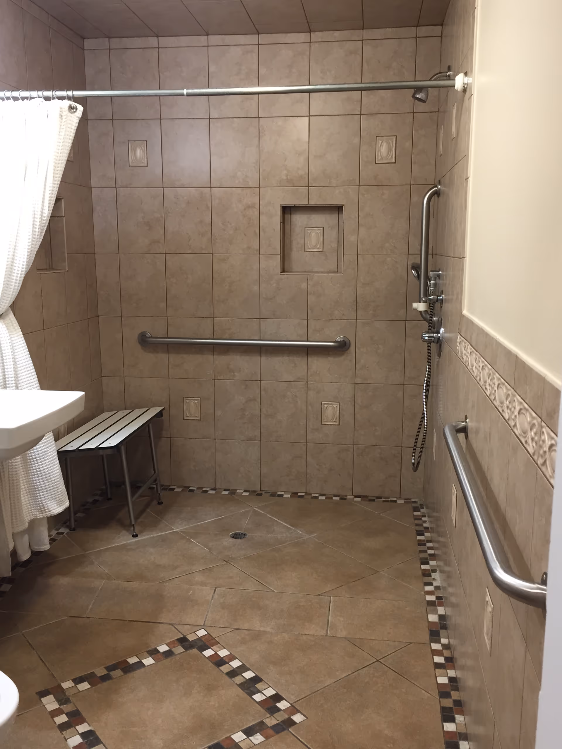 A tiled bathroom with beige and brown tiles on the walls and floor. The shower area has a metal grab bar, a handheld showerhead, and a small bench. There is a white shower curtain partially visible on the left side and a white sink also partially visible. The bathroom is designed for accessibility.