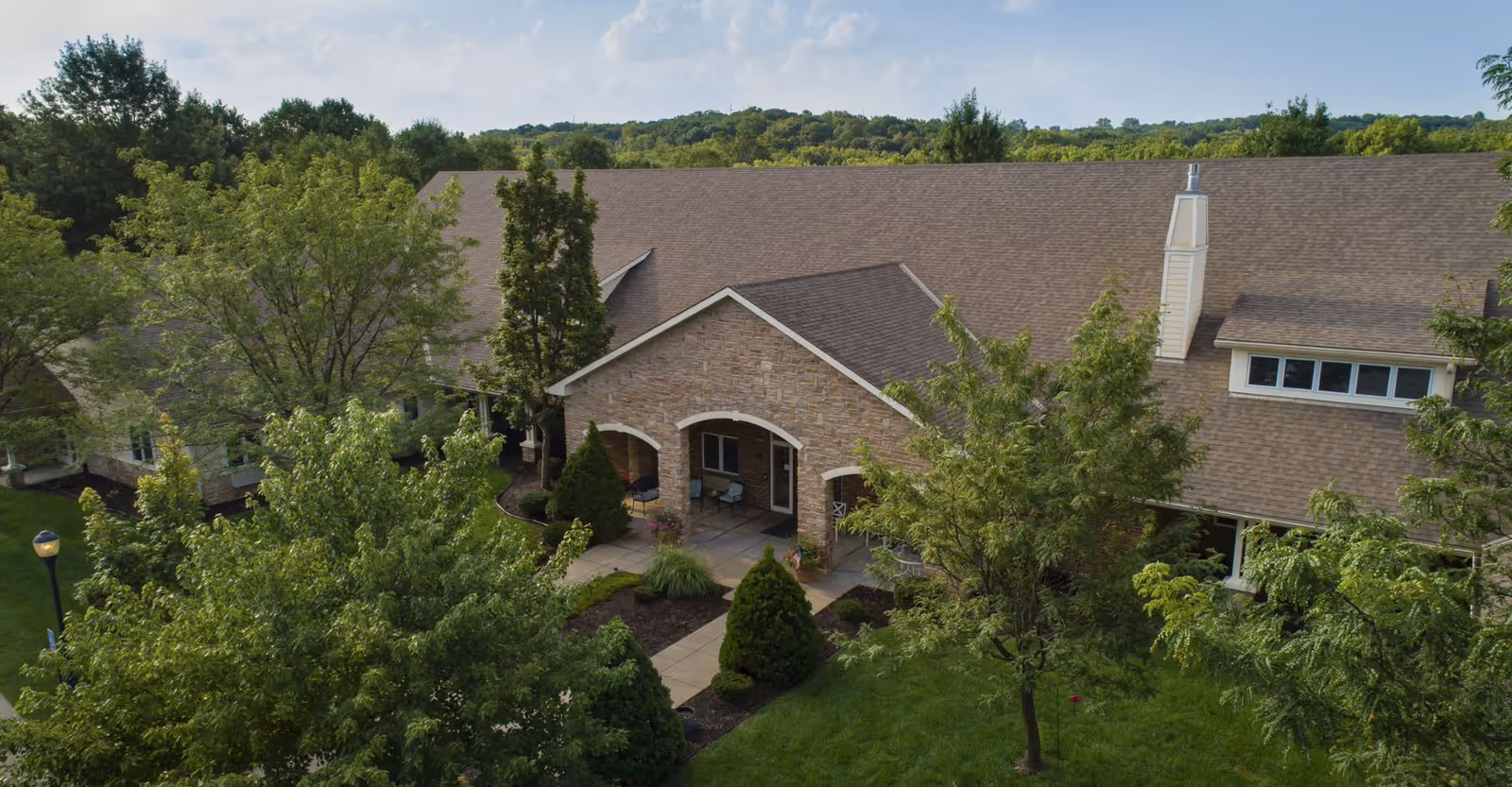 Aerial view of a single-story assisted living building with a stone arched entrance surrounded by trees and landscaped grounds.