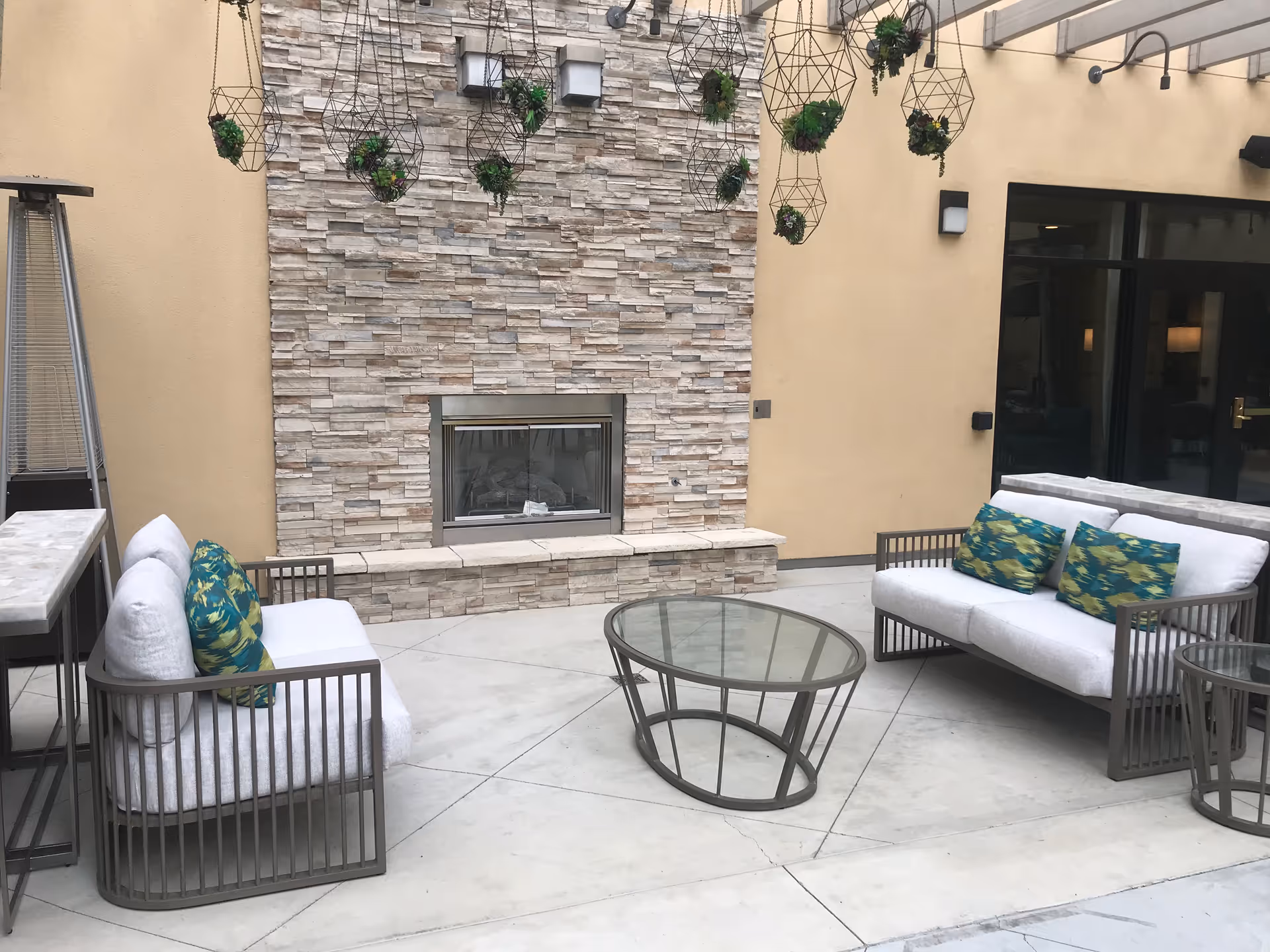Outdoor patio area with two cushioned sofas featuring green and yellow patterned pillows, a glass-top coffee table, and a stone fireplace built into a beige wall. Several hanging planters with green plants are suspended above the seating area.