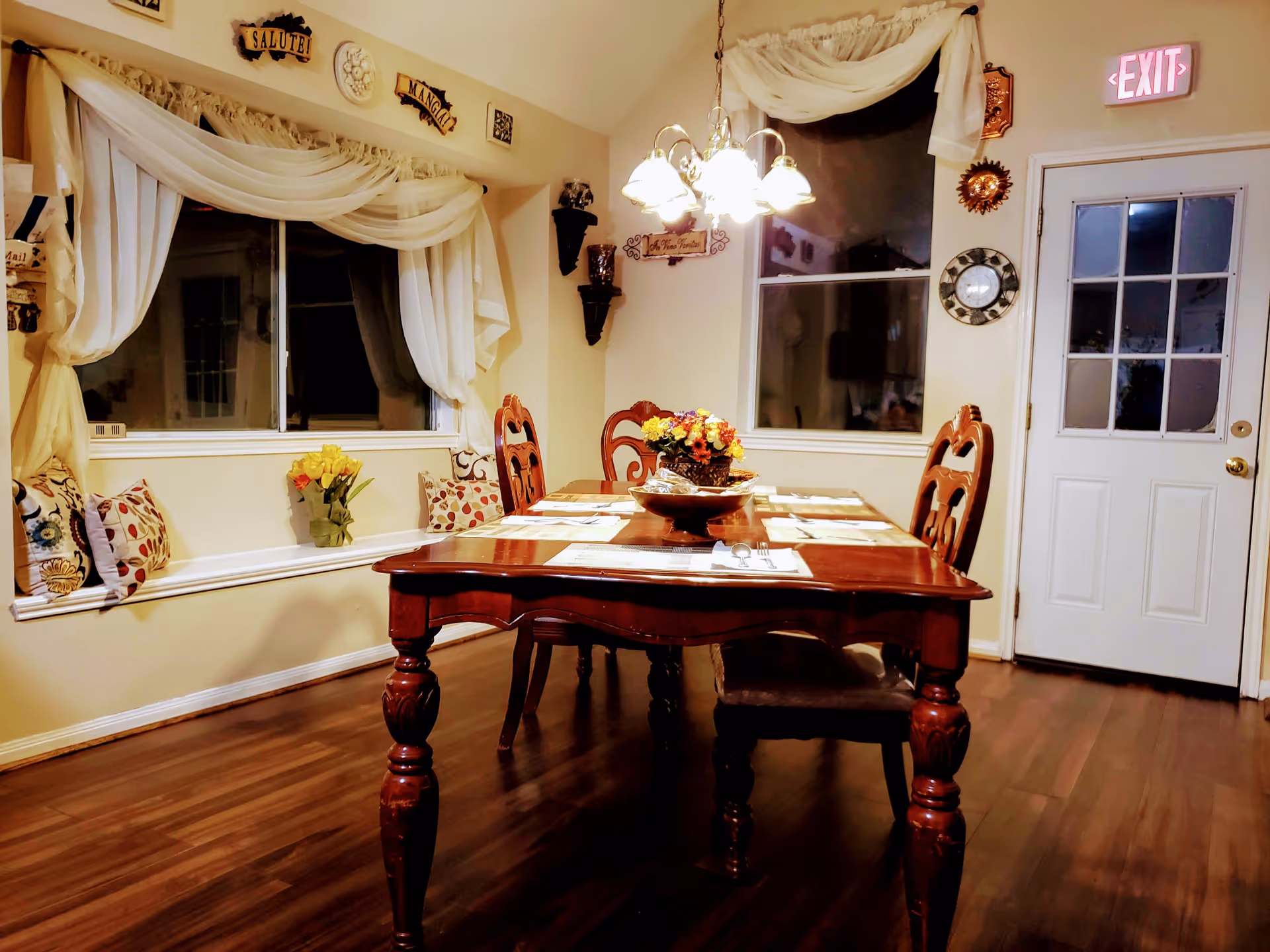 A warmly lit dining room with a wooden dining table set for four, featuring placemats and a centerpiece of flowers. The room has hardwood floors, two windows with white curtains, decorative pillows on a built-in bench under one window, and various wall decorations. A white door with glass panels and an illuminated exit sign is visible on the right side.