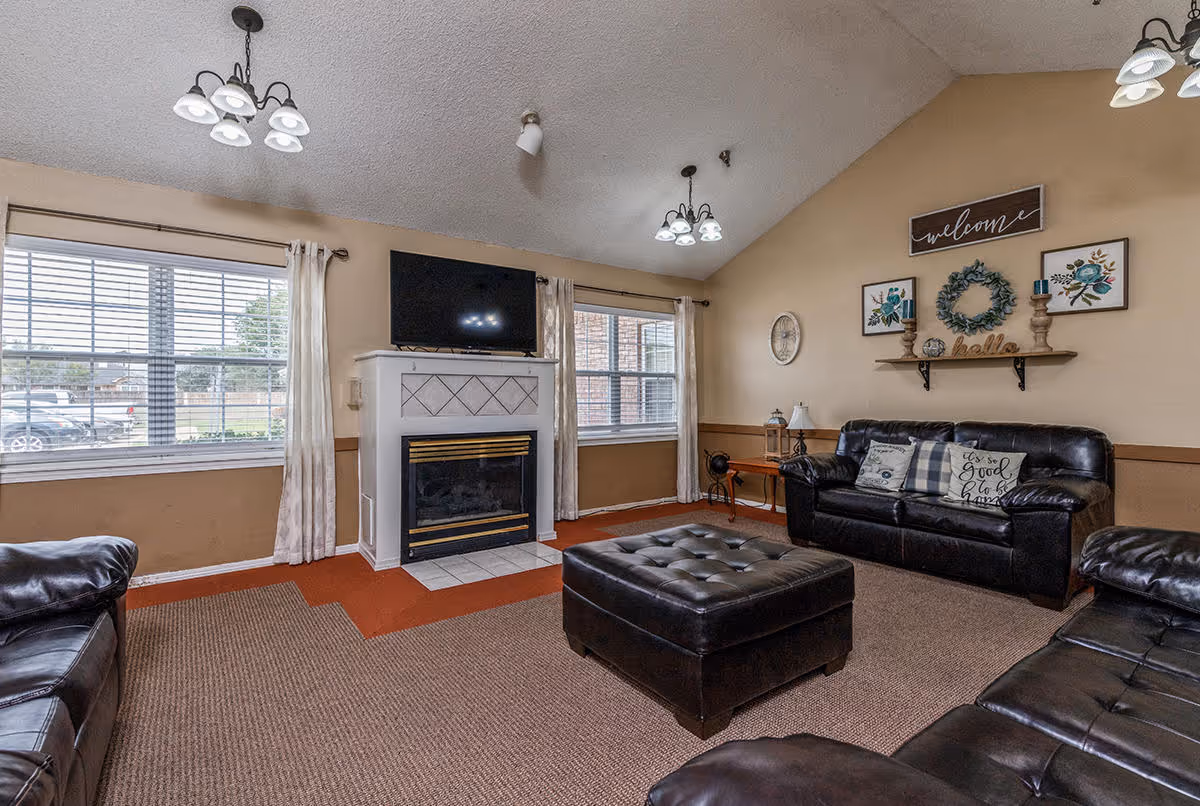 A cozy living room with beige walls and carpeted floor featuring three dark leather sofas arranged around a large tufted leather ottoman. A white fireplace with a mounted flat-screen TV above it is centered between two large windows with white curtains. The wall above one sofa is decorated with a 'welcome' sign, floral artwork, a wreath, and candles on a wooden shelf. Ceiling lights with multiple bulbs hang from the vaulted ceiling.