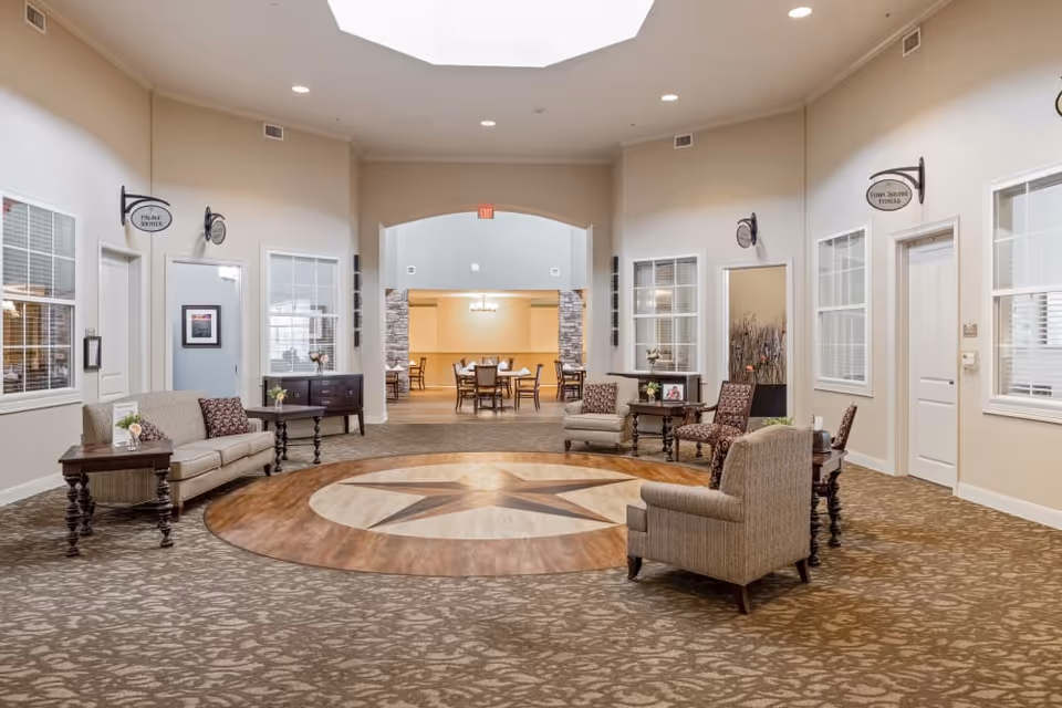 A spacious and well-lit common area in a senior living facility featuring a large circular wooden floor design with a star in the center. The room is furnished with several upholstered chairs and sofas arranged around small tables with flower vases. There are windows and doors along the walls, with signs indicating rooms such as the 'Pulse Center' and 'Town Square Dining'. In the background, a dining area with tables and chairs is visible through an arched doorway.