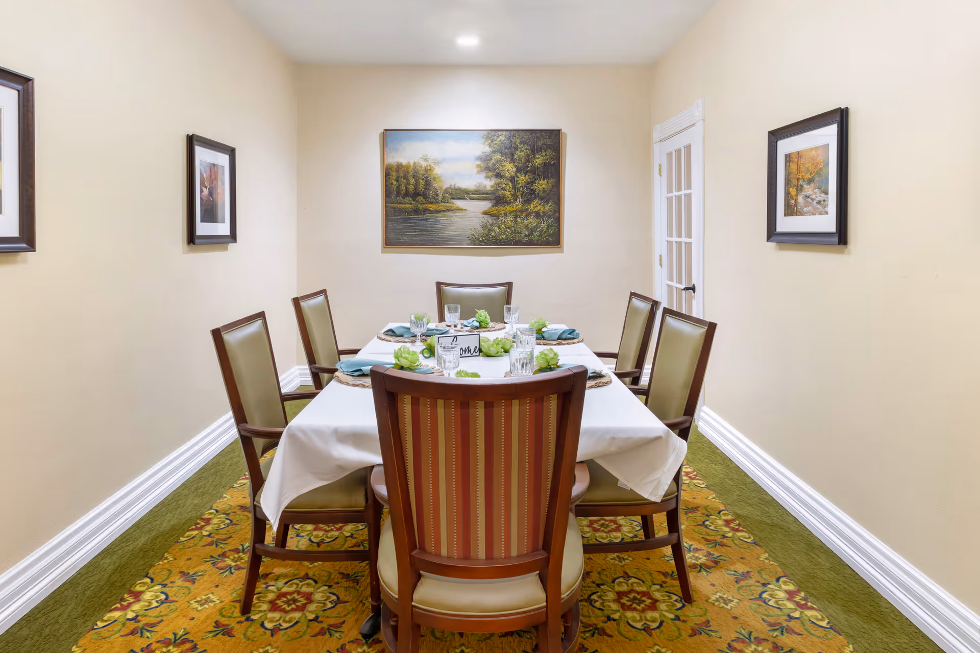 A small dining room with a rectangular table covered with a white tablecloth, set for six people with glasses, napkins, and decorative green plants. The room has beige walls adorned with three framed paintings, a patterned carpet with green and yellow tones, and a door with glass panels on the right side.