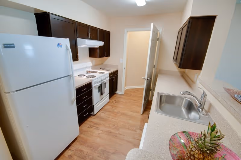 A clean galley kitchen with white appliances, dark wood cabinets, laminate countertops and a sink on the right.