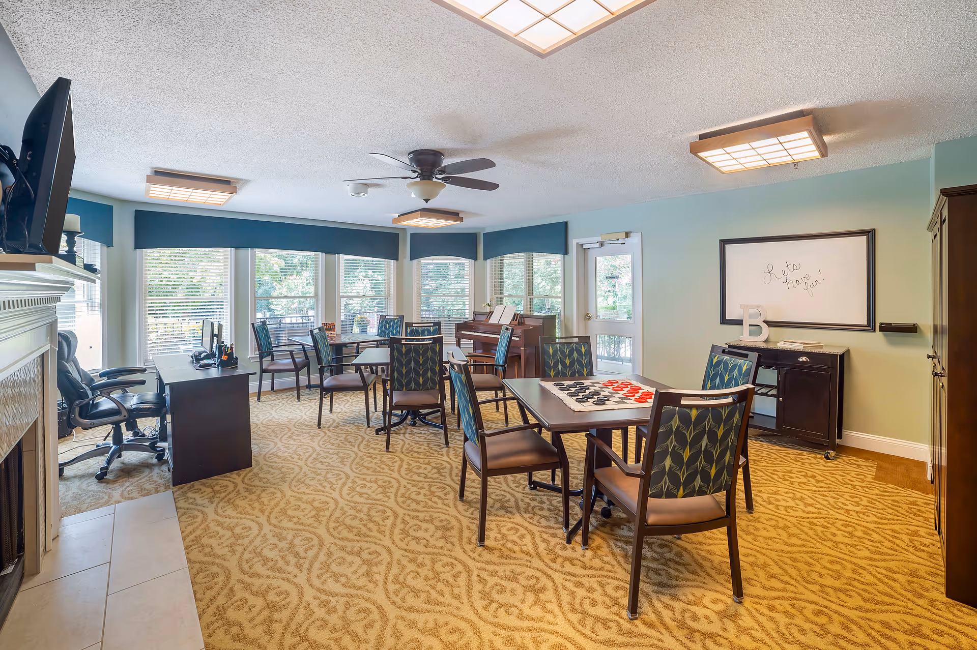 A bright and spacious common room with patterned carpet, several tables and chairs arranged for group activities, a piano near the windows, a ceiling fan, and a whiteboard on the wall with the message 'Let's have fun!'. Large windows allow natural light to fill the room, and there is a desk with office chairs near a fireplace on the left side.