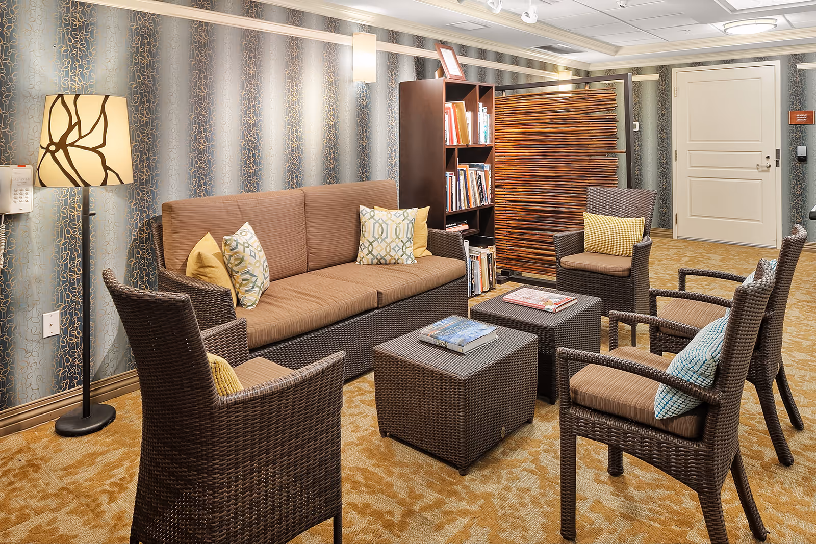 Cozy lounge with a wicker sofa and armchairs arranged around coffee tables, a bookshelf, and a floor lamp.