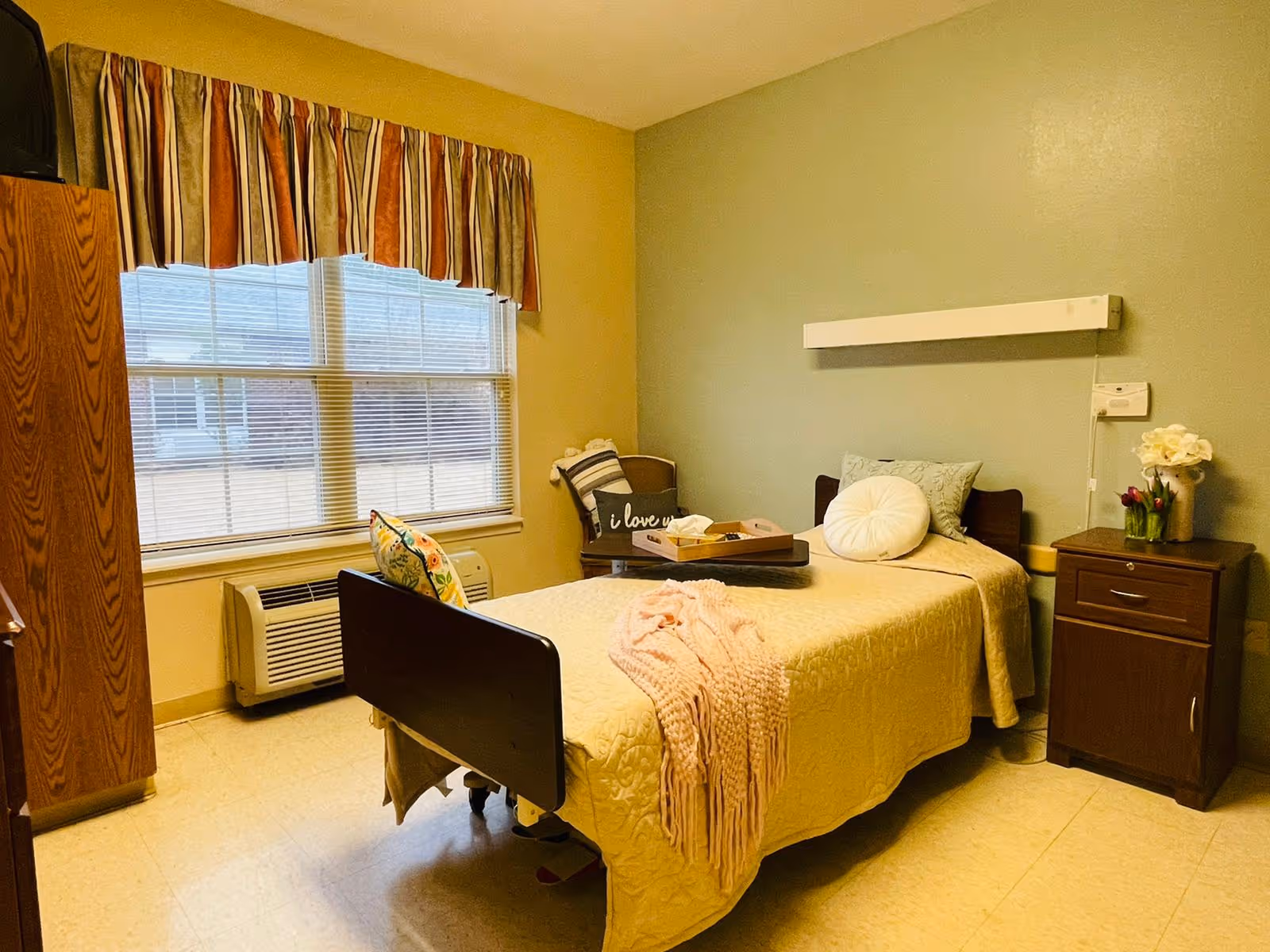 A single bed in a small, cozy bedroom with a yellow bedspread, a pink throw blanket, and decorative pillows. There is a wooden nightstand with flowers on it to the right of the bed, a chair with a cushion and a pillow that says 'I love u' in the corner, and a wooden cabinet with a TV on top to the left near a window with striped curtains. The walls are painted in two tones, yellow and light green.