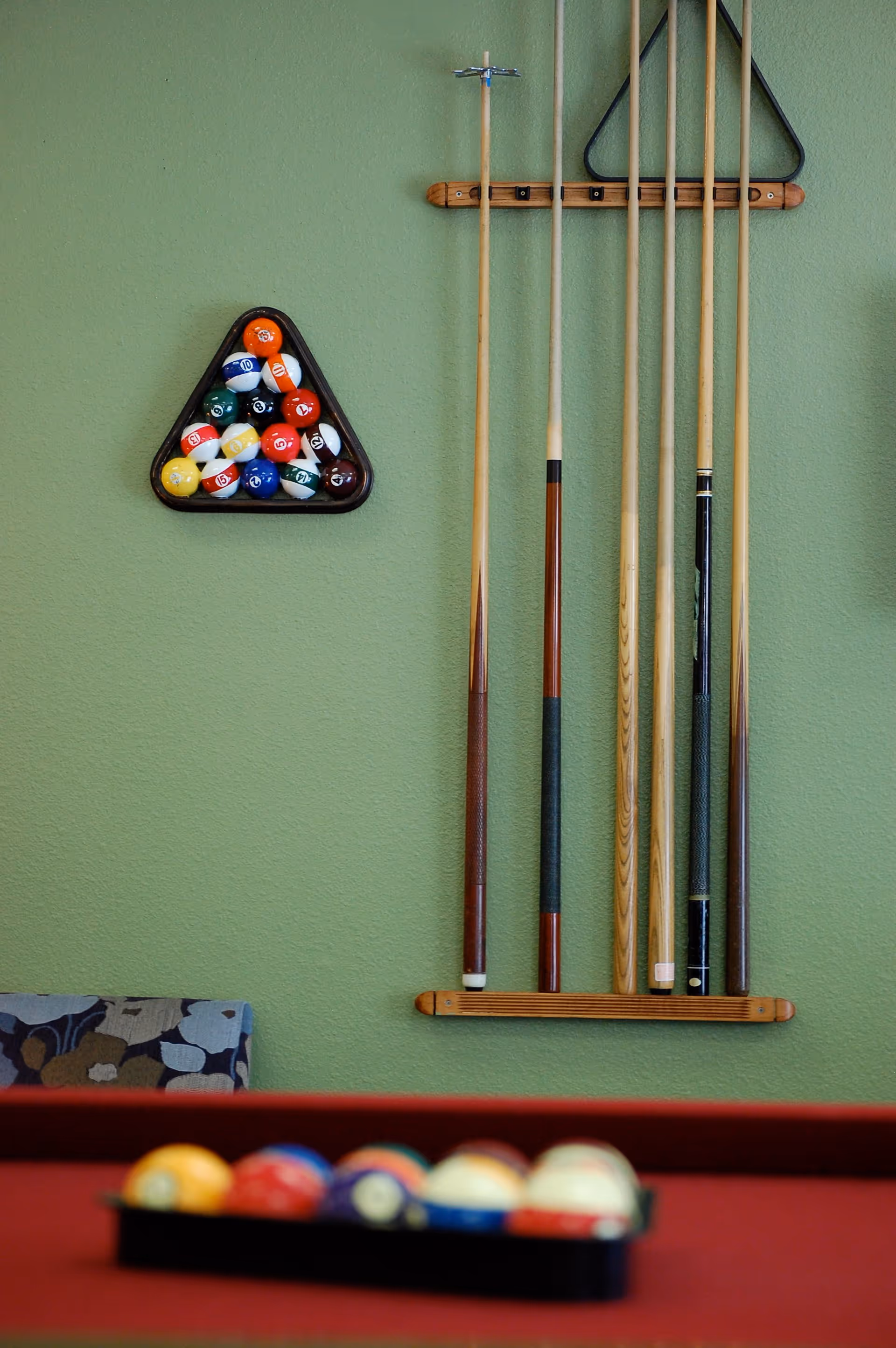 A green wall with a rack holding five pool cues and a triangular rack filled with billiard balls. In the foreground, a red pool table with a blurred rack of billiard balls is visible.