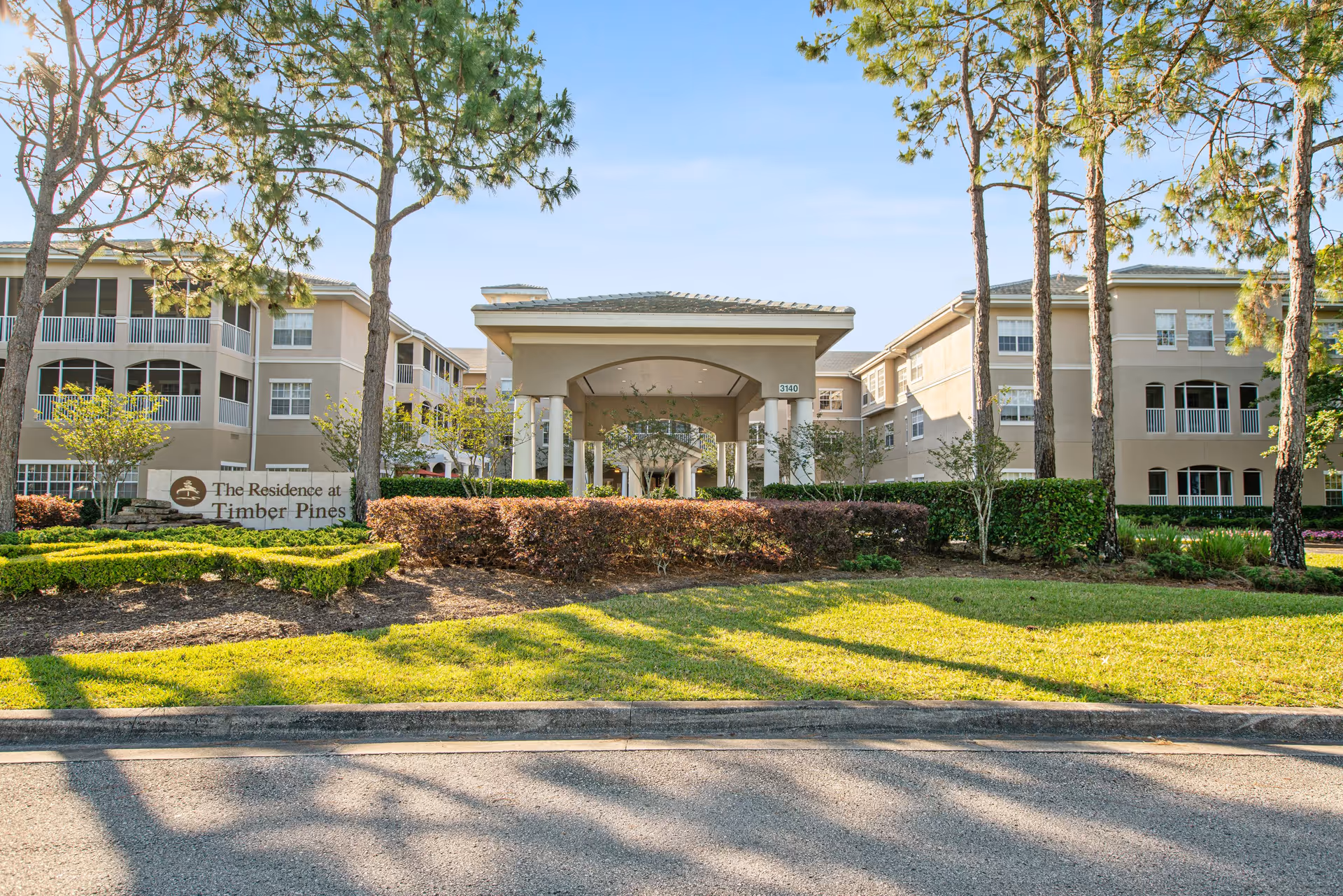 Exterior view of The Residence at Timber Pines senior living facility showing a large beige building with multiple windows and balconies, surrounded by trees, shrubs, and well-maintained landscaping under a clear blue sky.