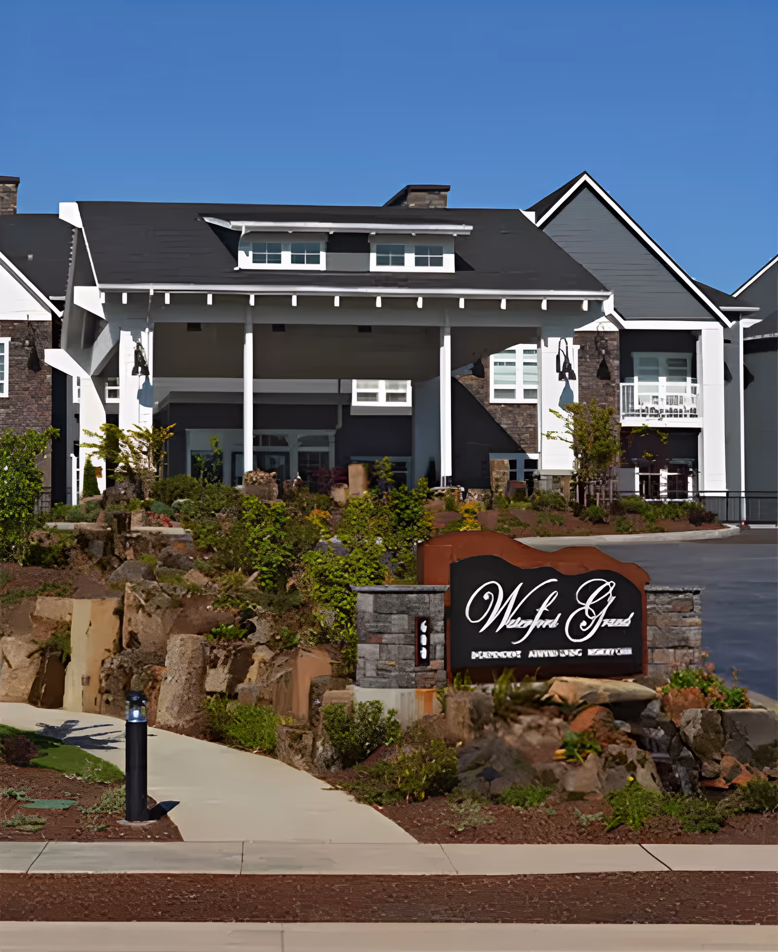 Front exterior view of Waterford Grand, a senior living facility, showing the main entrance with a covered driveway, landscaped garden with rocks and greenery, and a sign displaying the facility's name.