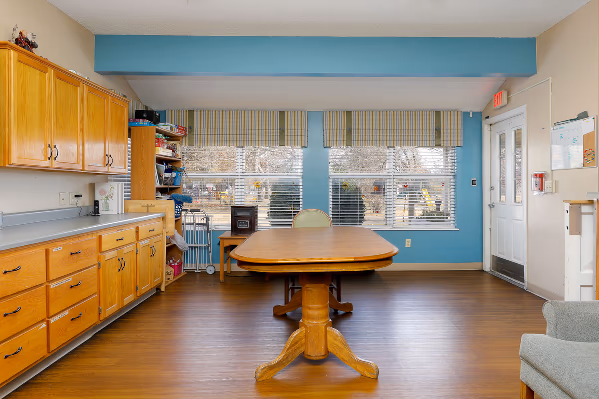 A room with wooden cabinets and drawers on the left side, a wooden table with a single chair in front of three large windows with striped valances, and a door on the right side. The floor is wooden, and the walls are painted blue and beige.