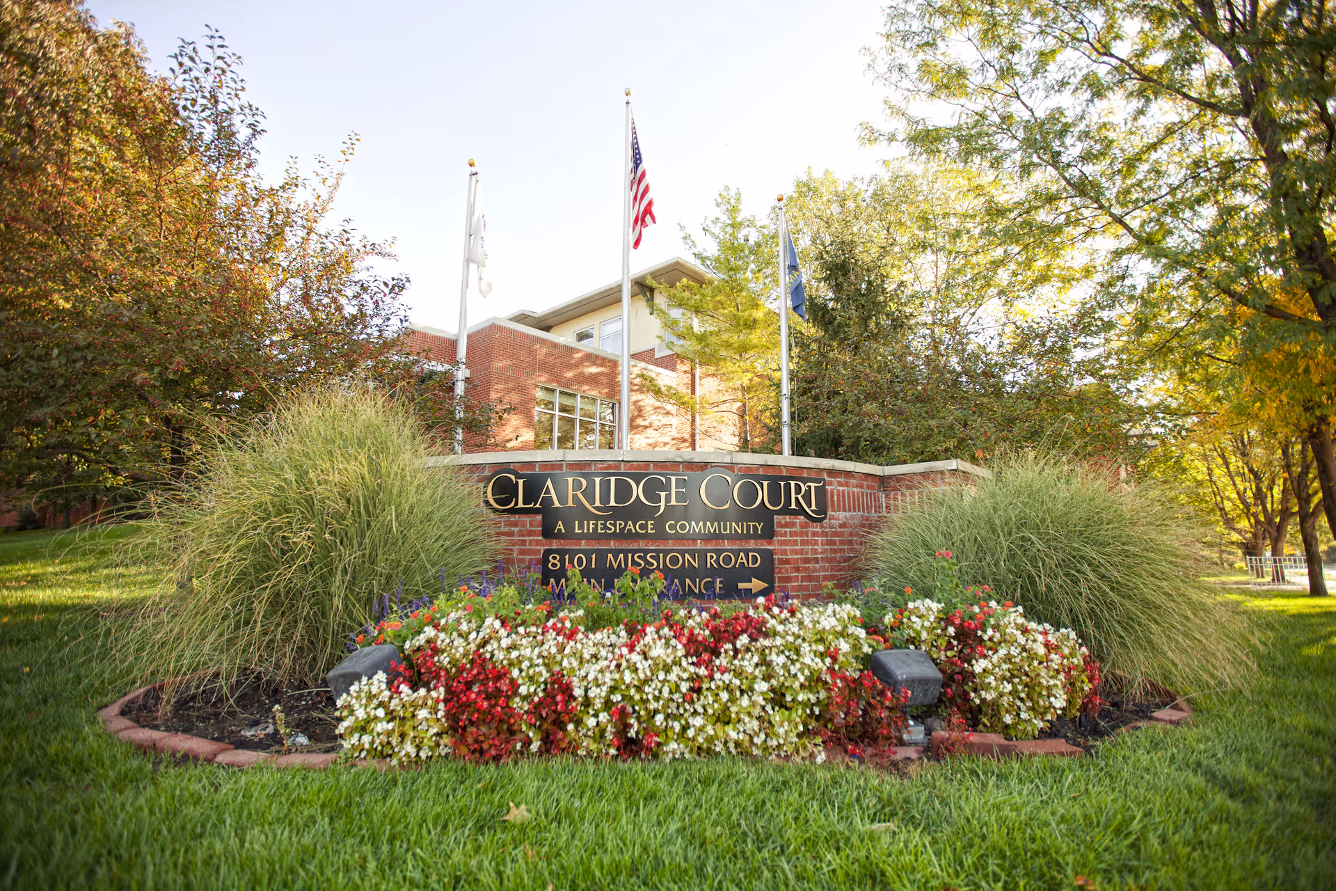Outdoor view of the Claridge Court senior living community sign surrounded by green grass, colorful flowers, and trees with a brick building and three flagpoles in the background.
