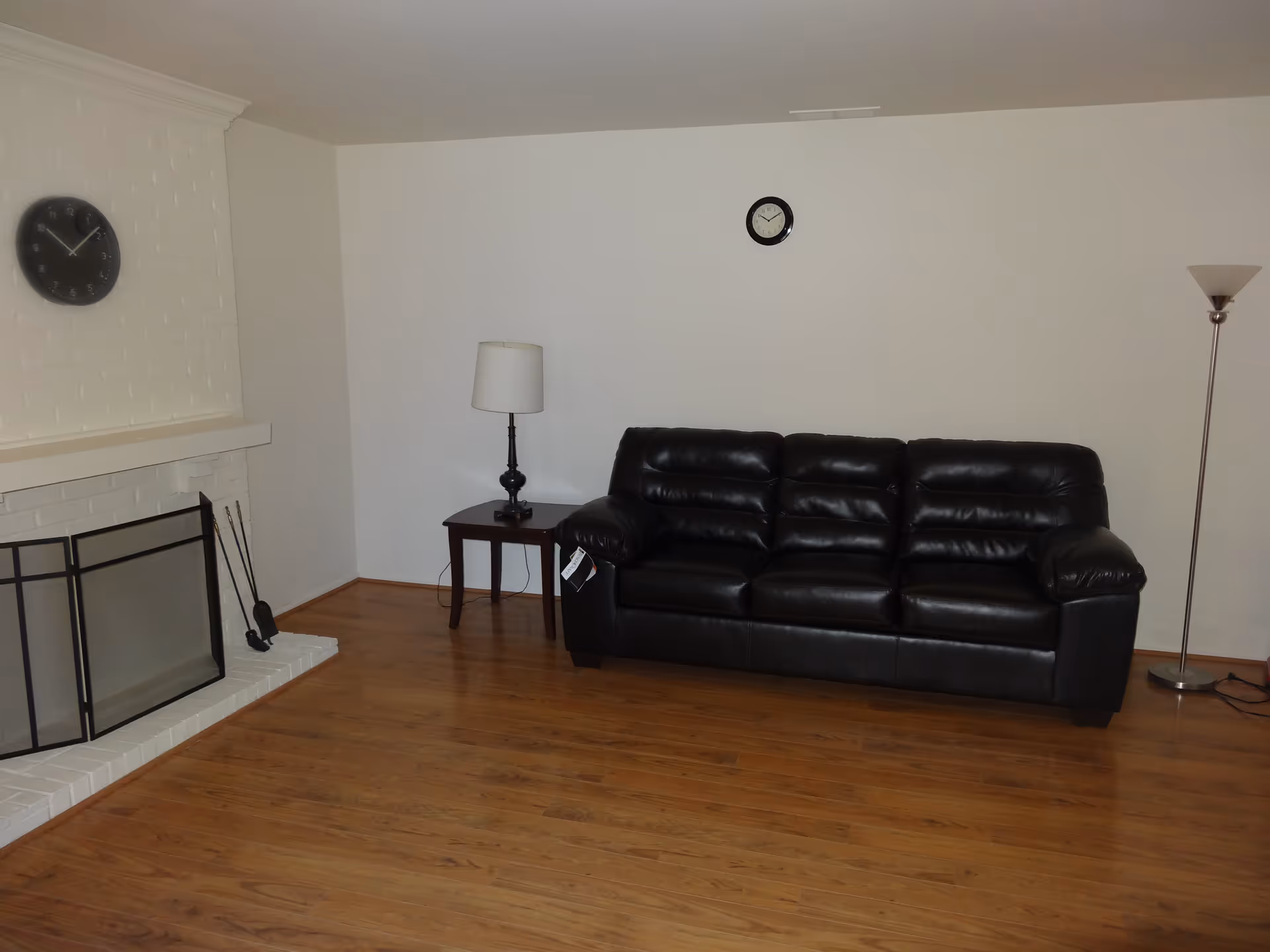 A living room with a black leather sofa, a small wooden side table with a lamp, a tall floor lamp, two wall clocks, and a white brick fireplace with a black screen and fireplace tools. The floor is wooden and the walls are plain white.