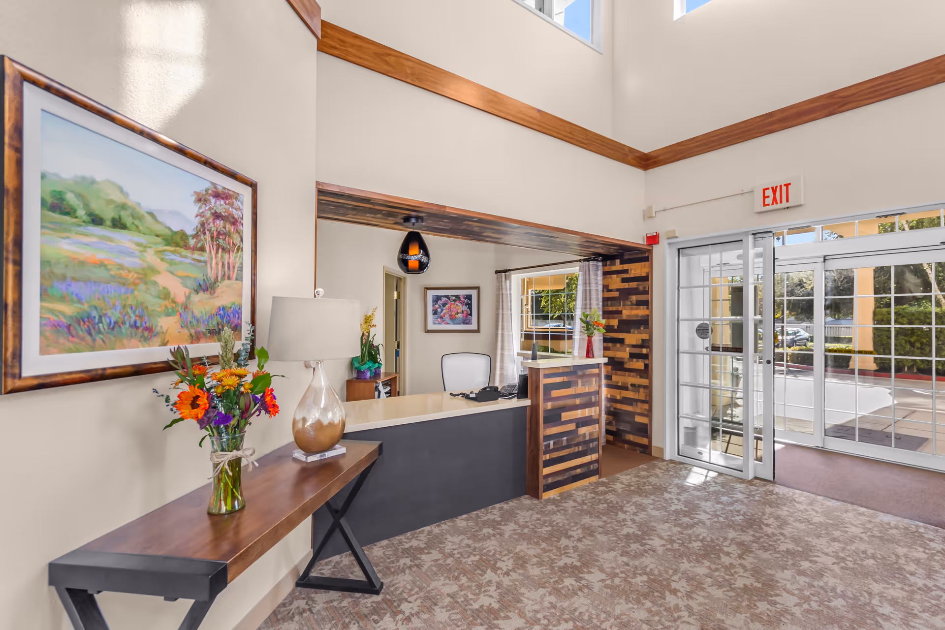 Reception area of Merrill Gardens At Santa Maria featuring a wooden desk with a chair behind it, a table with a lamp and a vase of colorful flowers, a large framed landscape painting on the wall, and glass double doors leading outside with an exit sign above.