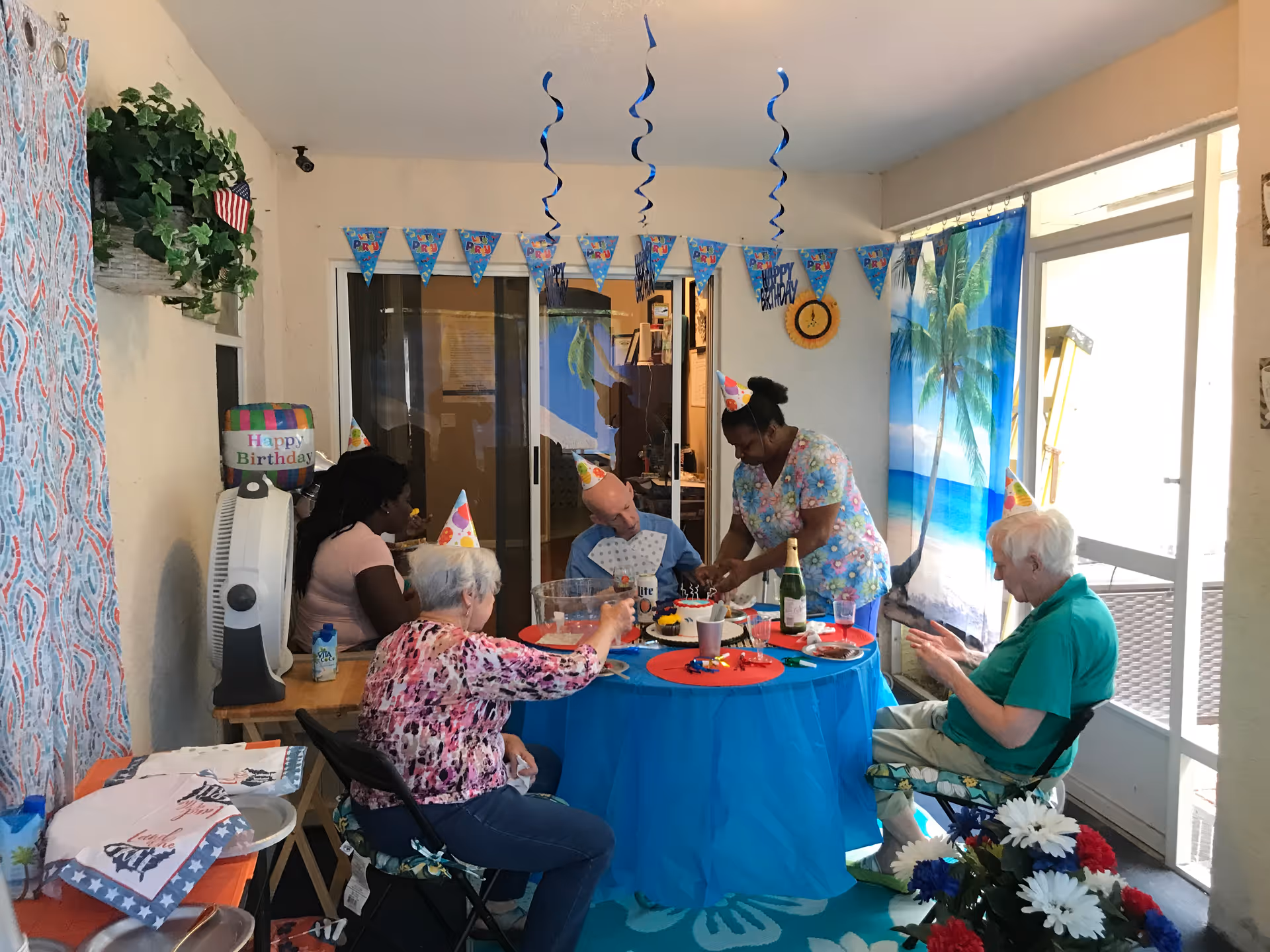 A group of elderly people and a caregiver wearing party hats celebrating a birthday around a table decorated with a blue tablecloth, birthday cake, and party decorations in a residential care facility room. The room has a tropical beach curtain, hanging blue streamers, and a Happy Birthday banner.