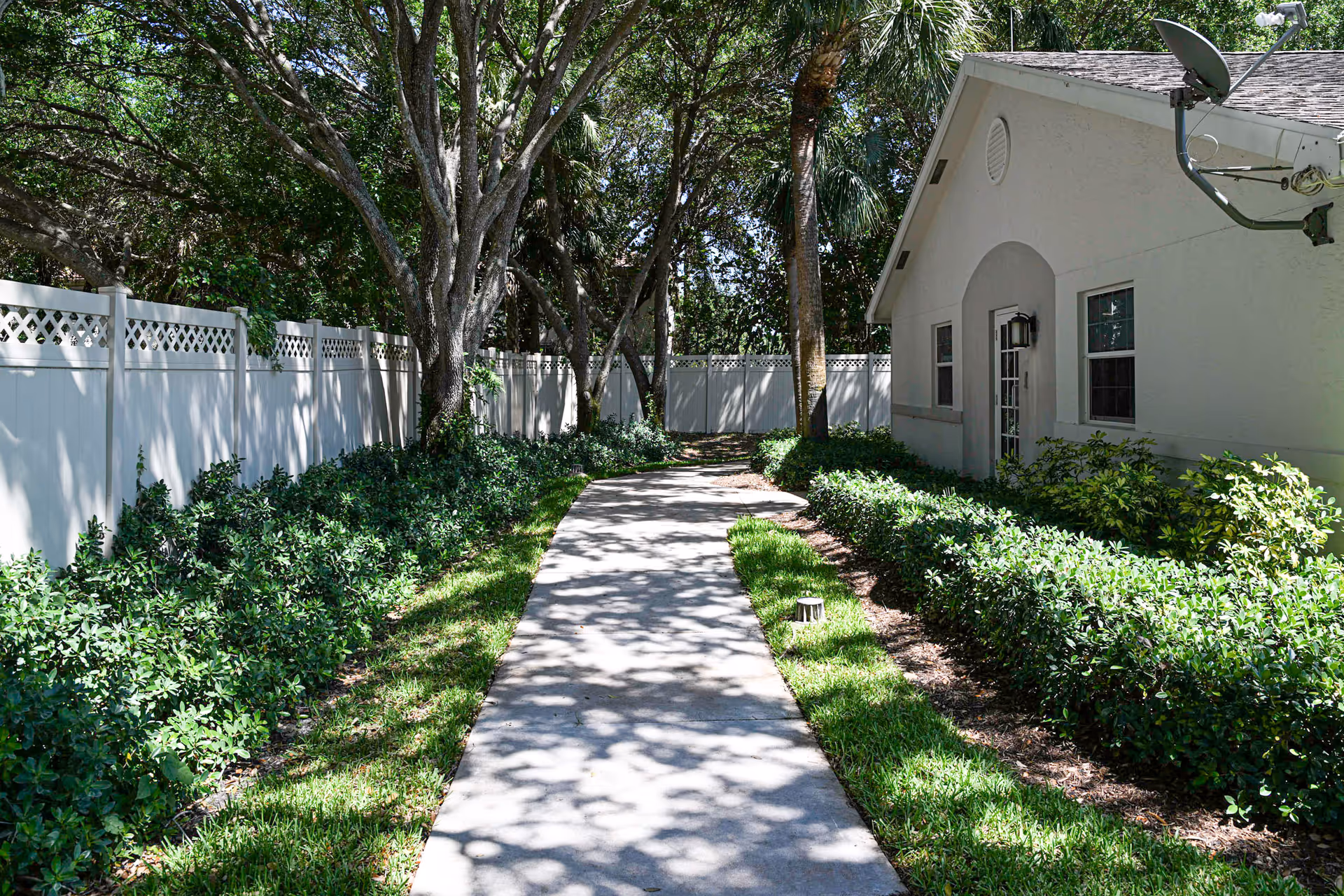 A concrete walkway flanked by green bushes and grass leads to a wooded area. On the right side, there is a white building with windows, a door, and a satellite dish on the roof. On the left side, there is a white fence with lattice design at the top. Trees cast shadows on the walkway and surrounding greenery.