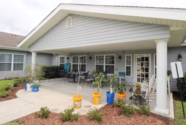 Covered outdoor patio area at a senior living facility with several chairs and tables, potted plants in colorful buckets, and a garden bed with small shrubs in front.