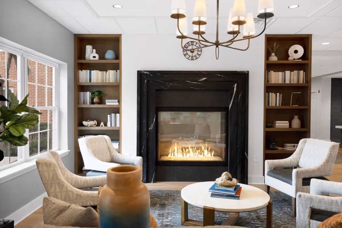A cozy living room area with a modern black marble fireplace in the center, surrounded by built-in wooden bookshelves filled with books and decorative items. Four upholstered armchairs are arranged around a round white coffee table with books and decorative objects on it. A large window on the left lets in natural light, and a chandelier with multiple lights hangs from the ceiling.